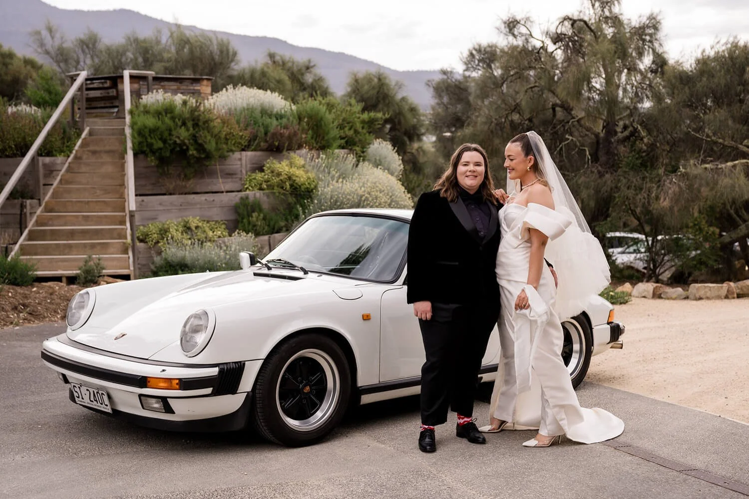 Two women standing next to a white vintage Porsche 911 on a paved surface, with a landscaped area and trees in the background. One woman is dressed in a black suit, and the other in a white wedding gown with a veil, smiling and posing together.