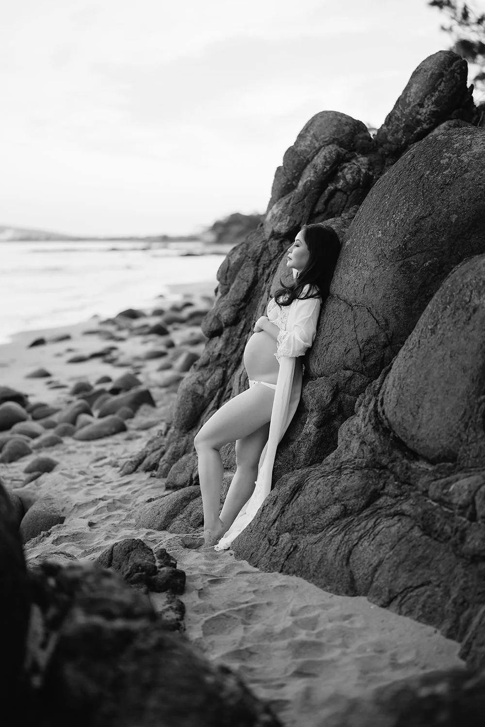 A pregnant woman with long dark hair stands on a sandy beach, leaning against a large rock formation with her eyes closed. She is wearing a sheer, long-sleeved white garment that reveals her pregnant belly.