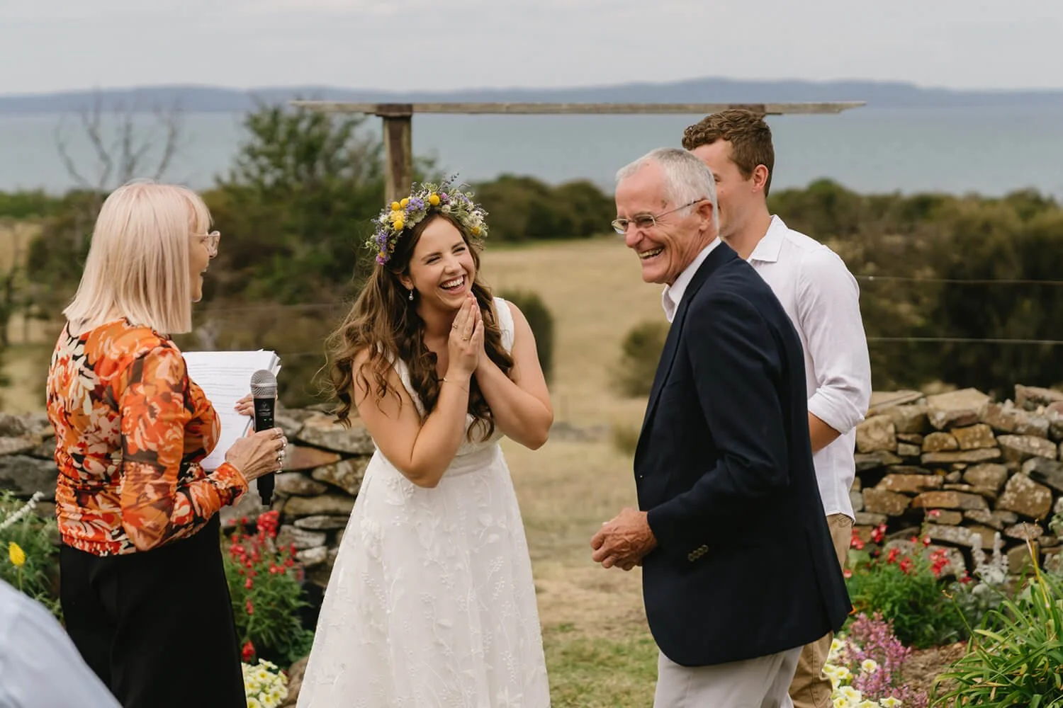 A bride smiling excitedly during her outdoor wedding ceremony with an officiant and guests, overlooking a scenic landscape with the water in the background.
