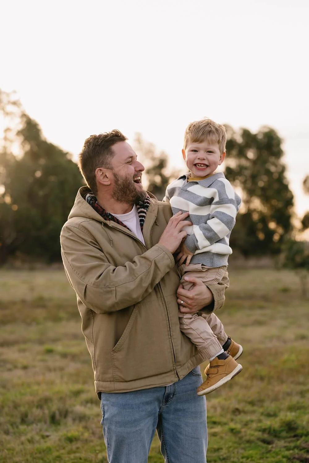 A man holding a smiling young boy outdoors at sunset, with trees in the background.