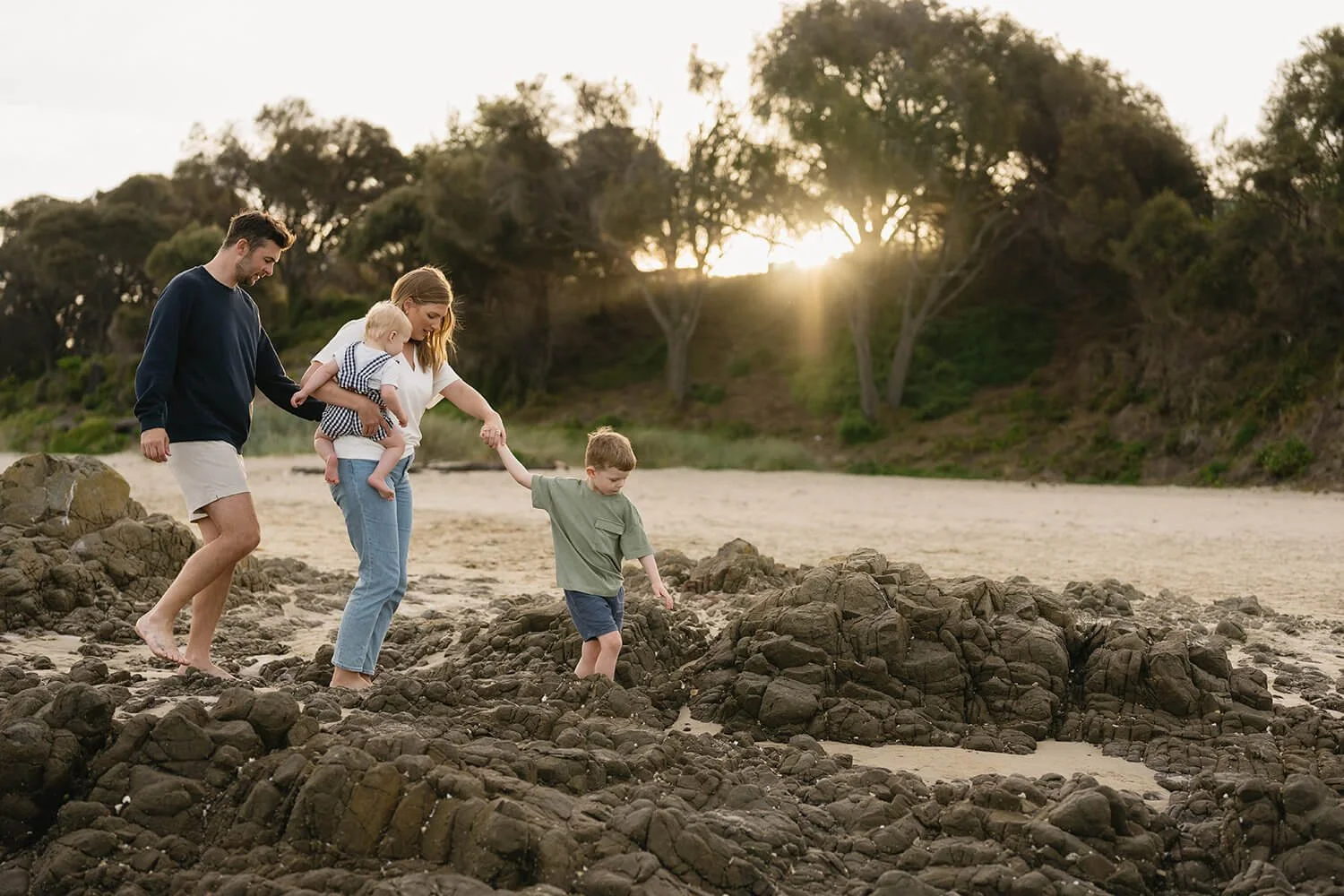 A family of four exploring rocks on a sandy beach during sunset. The father, mother, and two children walk barefoot on the rocks, with the children holding hands. The background features trees and the sun setting behind them.