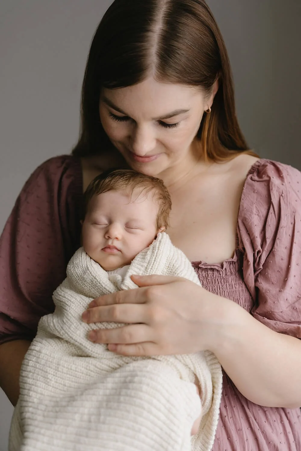A woman with shoulder-length brown hair and a pink dress holding a sleeping newborn wrapped in a cream-colored blanket against a plain gray backdrop.