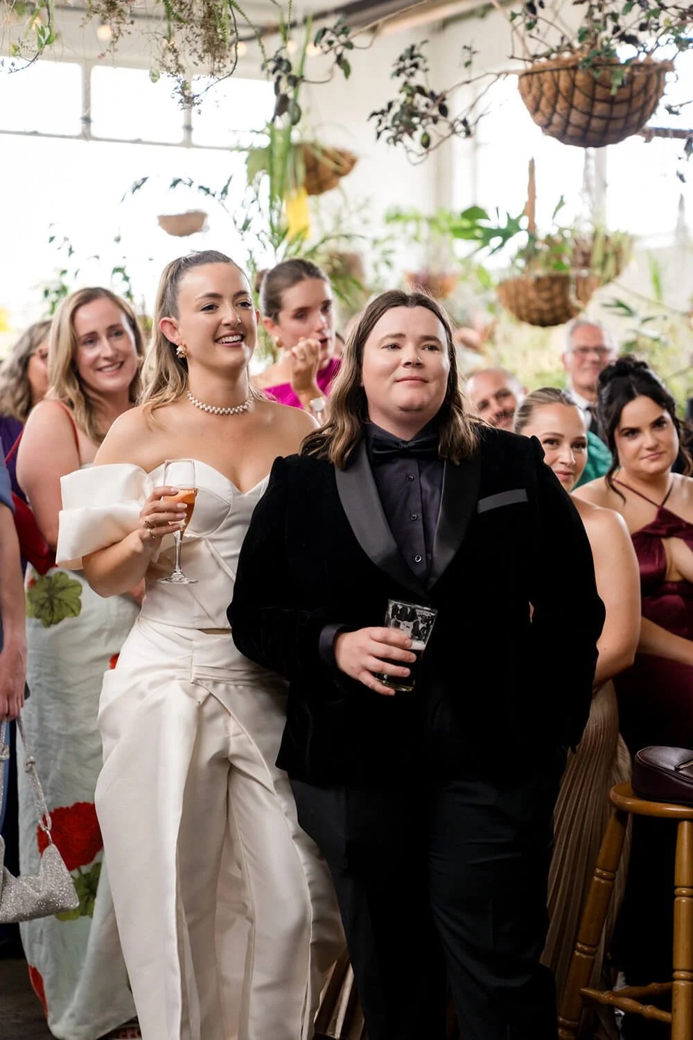 A group of people at a party, with a woman in a white dress holding a glass of champagne, smiling, standing next to a person in a black tuxedo holding a glass of beer, in a bright room with hanging plants and a crowd in the background.