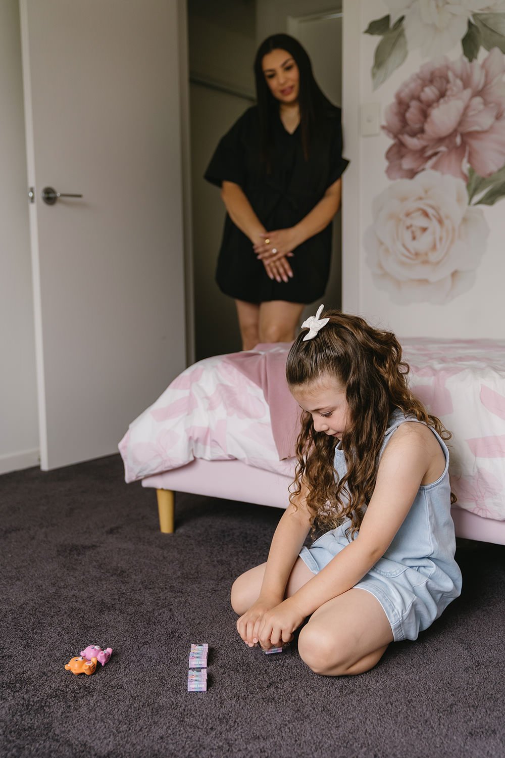 A young girl with curly hair, wearing a light blue sleeveless romper and a white hair bow, is sitting on a dark carpeted floor, playing with small toy pigs and a deck of cards. In the background, a woman with long black hair, dressed in a black outfi