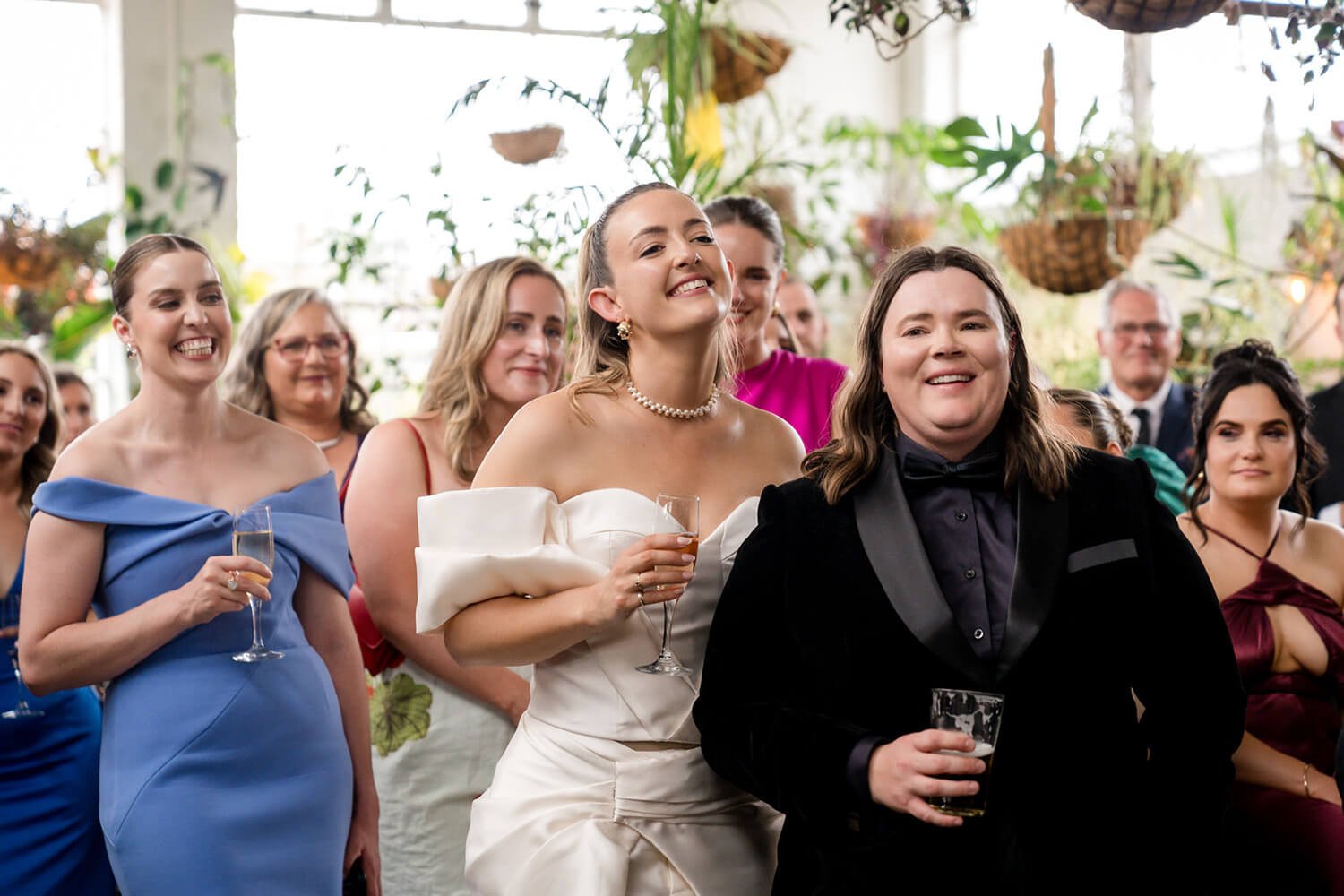People at a wedding reception, some holding drinks, smiling and looking in the same direction inside a decorated venue with hanging plants.