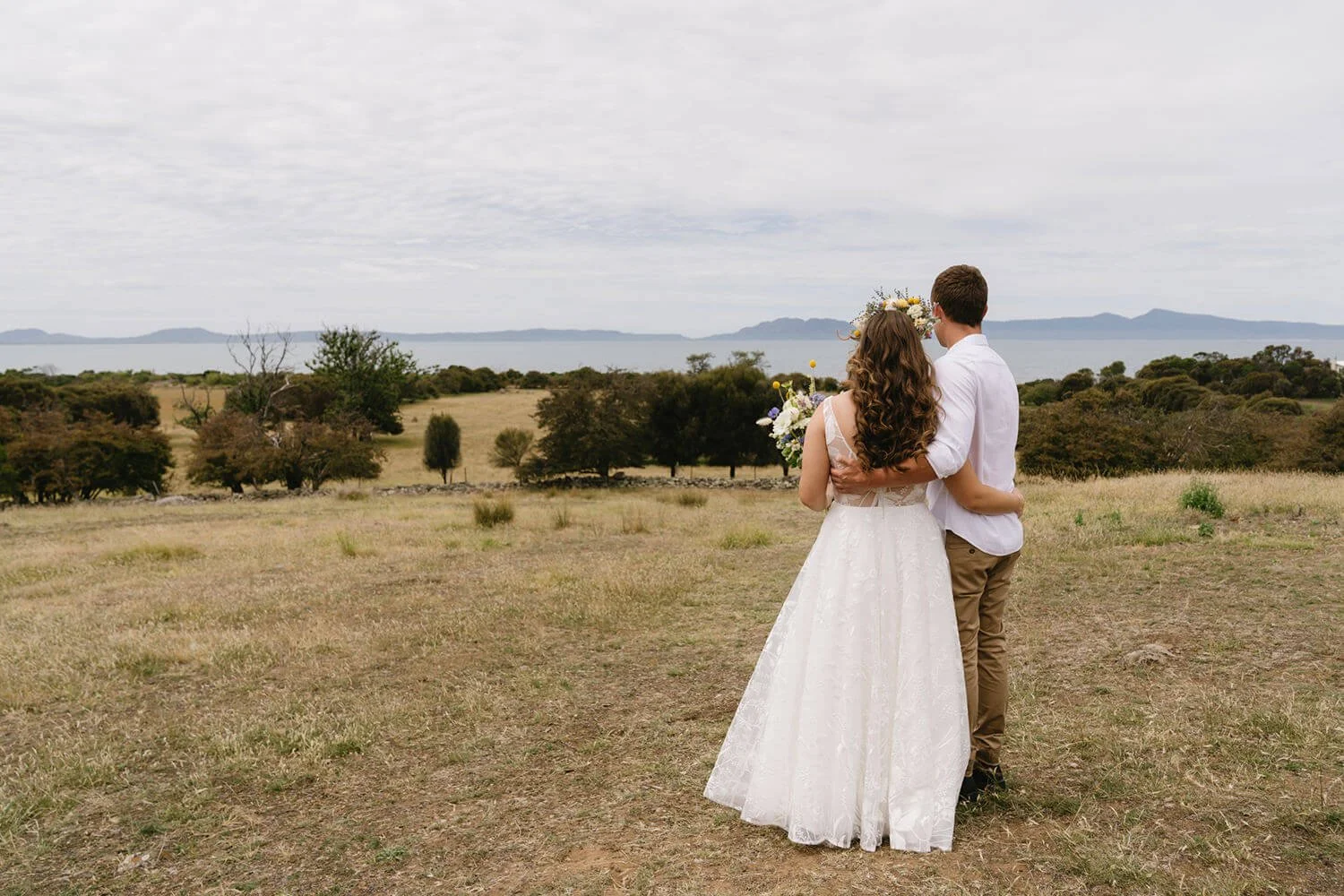 A couple on their wedding day standing outdoors in a field, embracing, with a scenic view of trees and distant mountains in the background, a cloudy sky overhead.