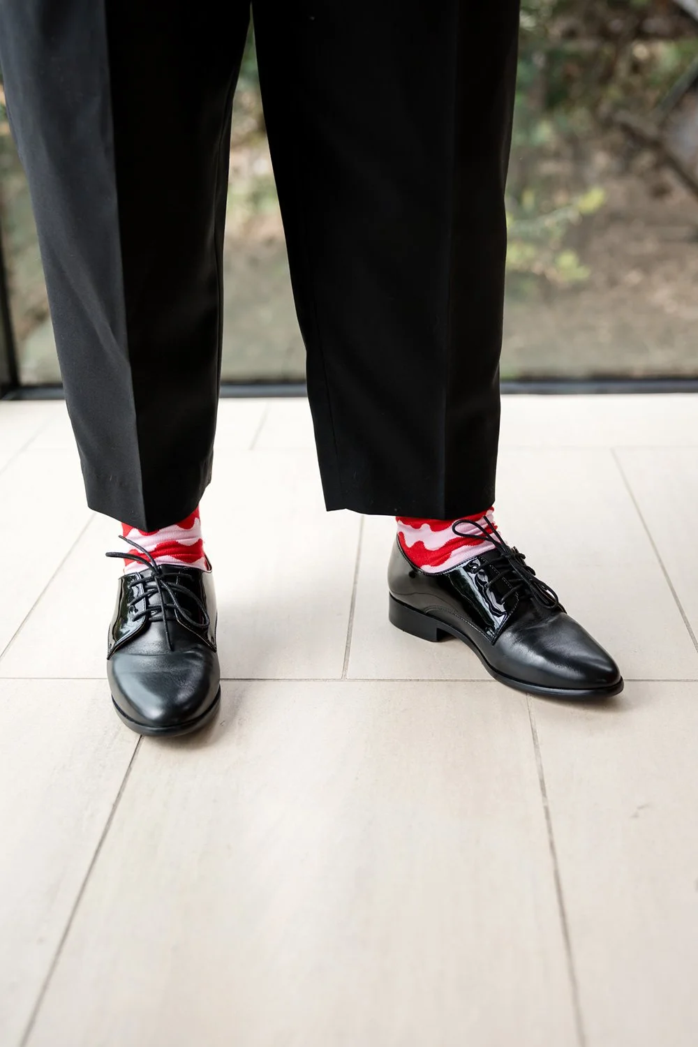 Person wearing black dress shoes, red and white patterned socks, black pants, standing on a tile floor with a window in the background.