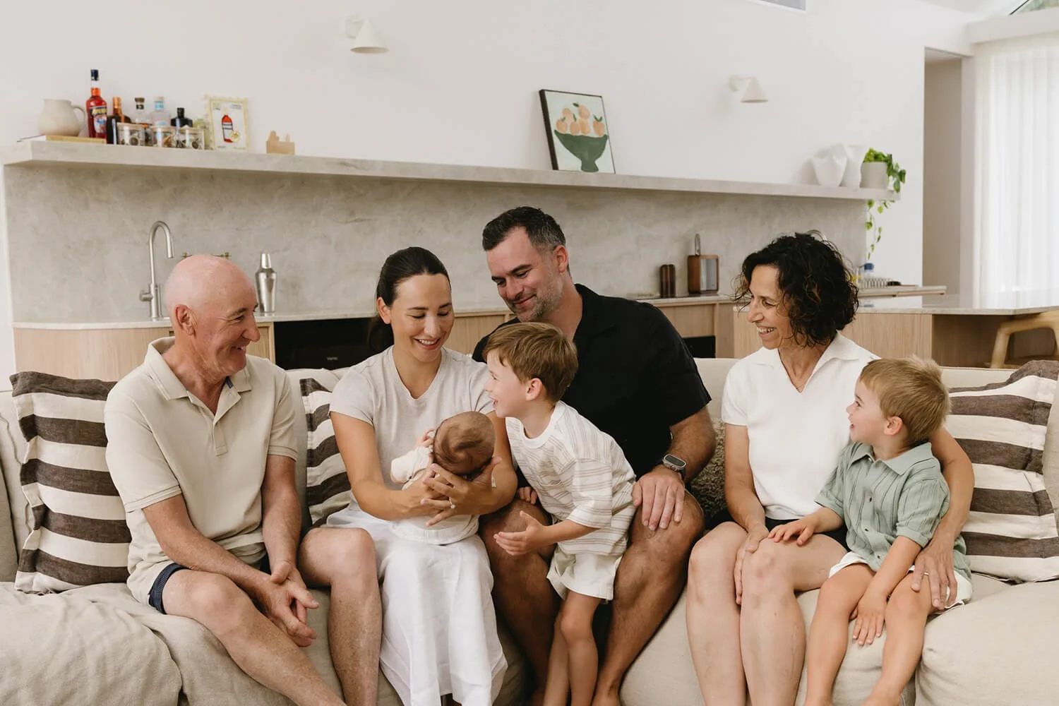 Family of seven sitting on a sofa in a bright living room, smiling and interacting with a newborn baby.