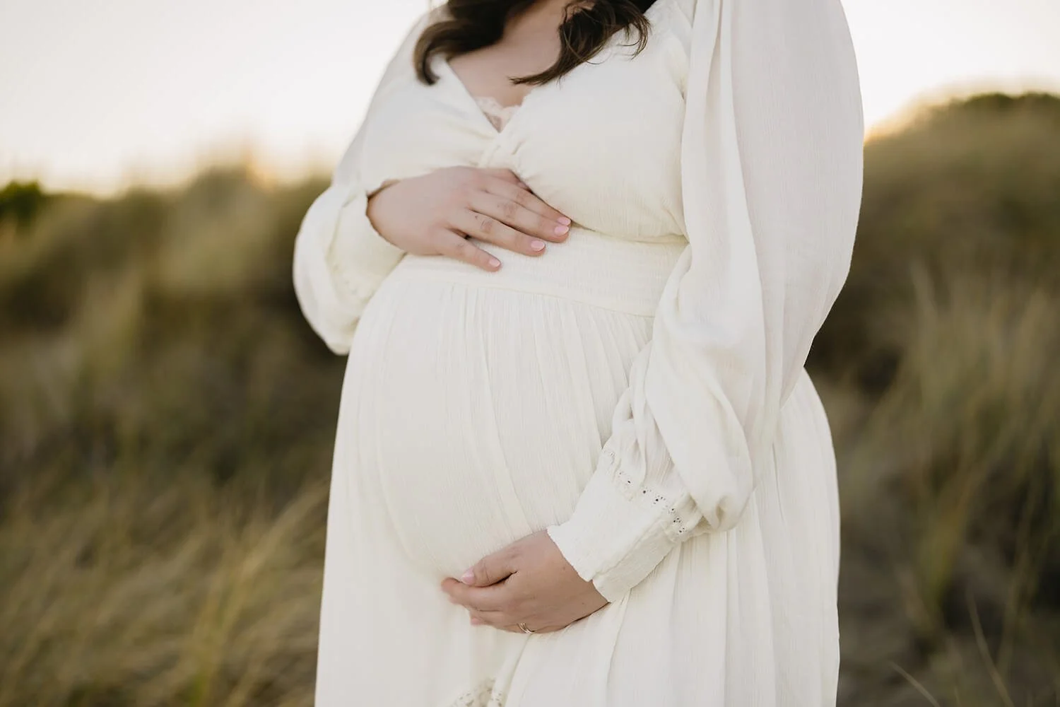 Close-up of a pregnant woman in a cream-colored dress holding her belly with one hand and her breast with the other, standing outdoors with a blurred natural background.
