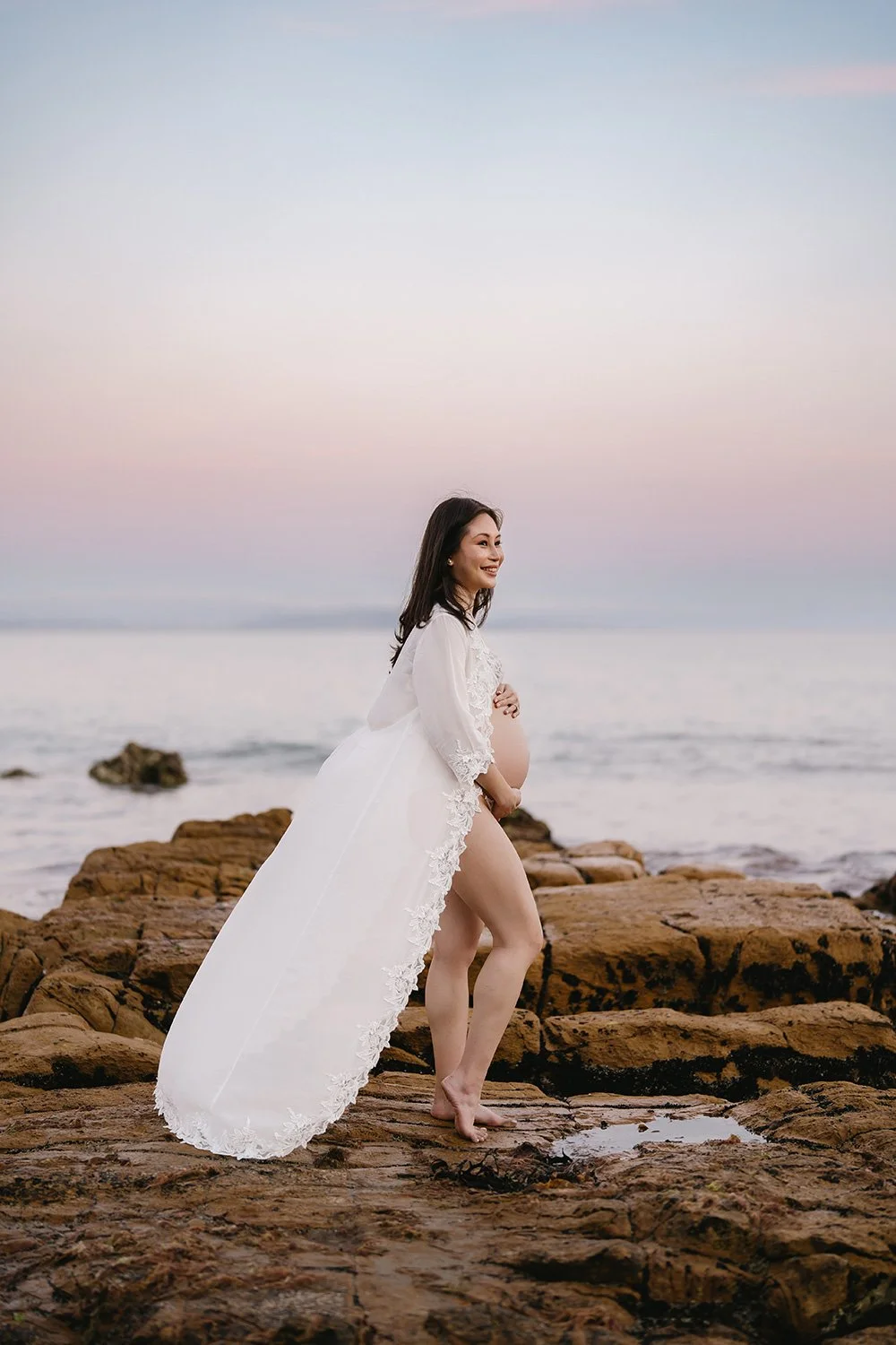 A pregnant woman in a white lace dress standing barefoot on rocks by the ocean during sunset, smiling and looking to the side.