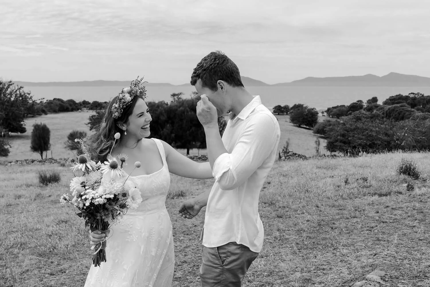 A woman in a wedding dress holding a bouquet, smiling and laughing while a man in casual clothes wipes away tears or wipes his eyes, standing outdoors in a scenic landscape with trees and mountains in the background.