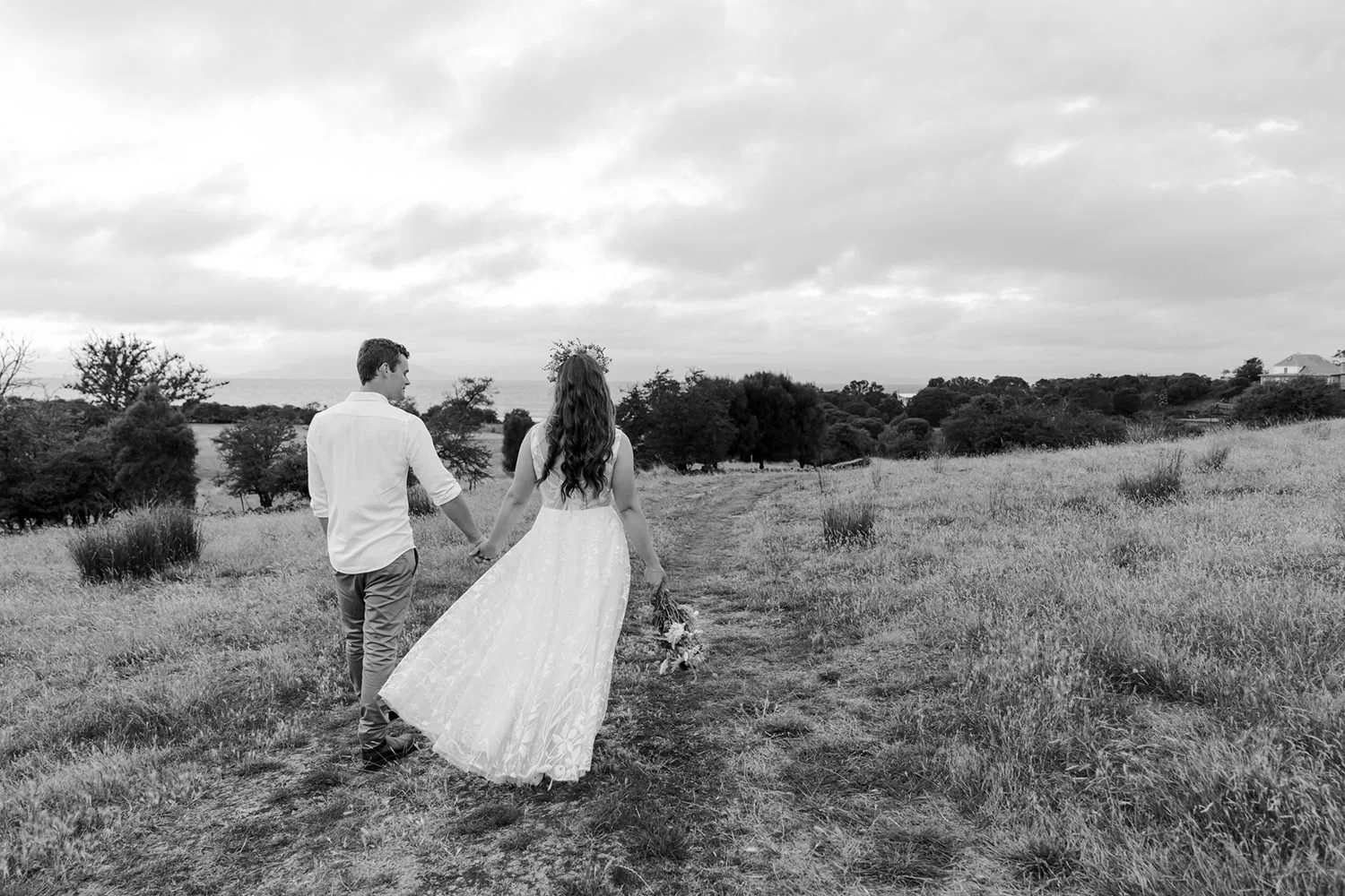 Black and white photo of a couple walking hand in hand along a grassy path in an open field with trees in the background. The woman is wearing a long dress and holding a bouquet, and the man is in casual attire.