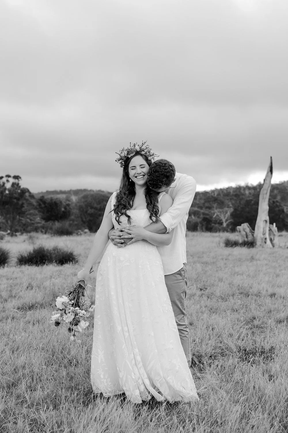 A joyful bride in a wedding dress with a floral crown, holding a bouquet, is being embraced from behind by a smiling groom in an outdoor field with trees and a cloudy sky in the background.