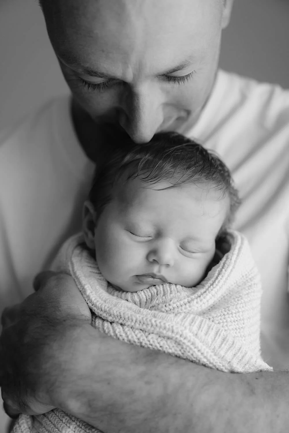 A man gently cradling a sleeping baby wrapped in a knitted blanket, with the man kissing the baby's forehead, in black and white.