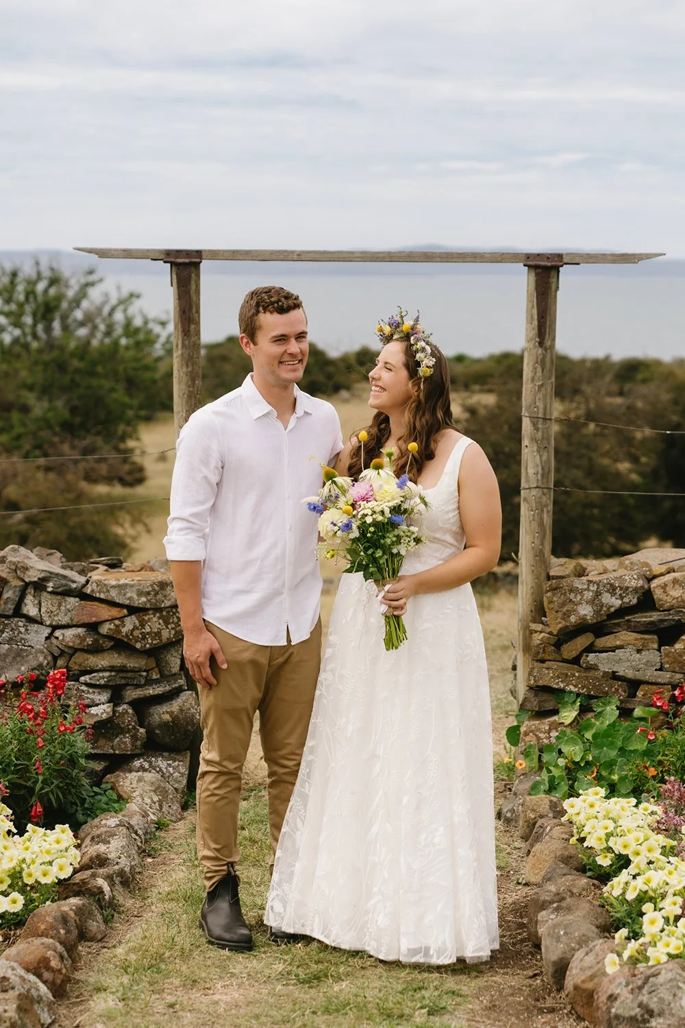 A smiling couple standing outdoors behind a rustic wooden arch, with stone walls and colorful flowers, during a wedding ceremony or photoshoot.