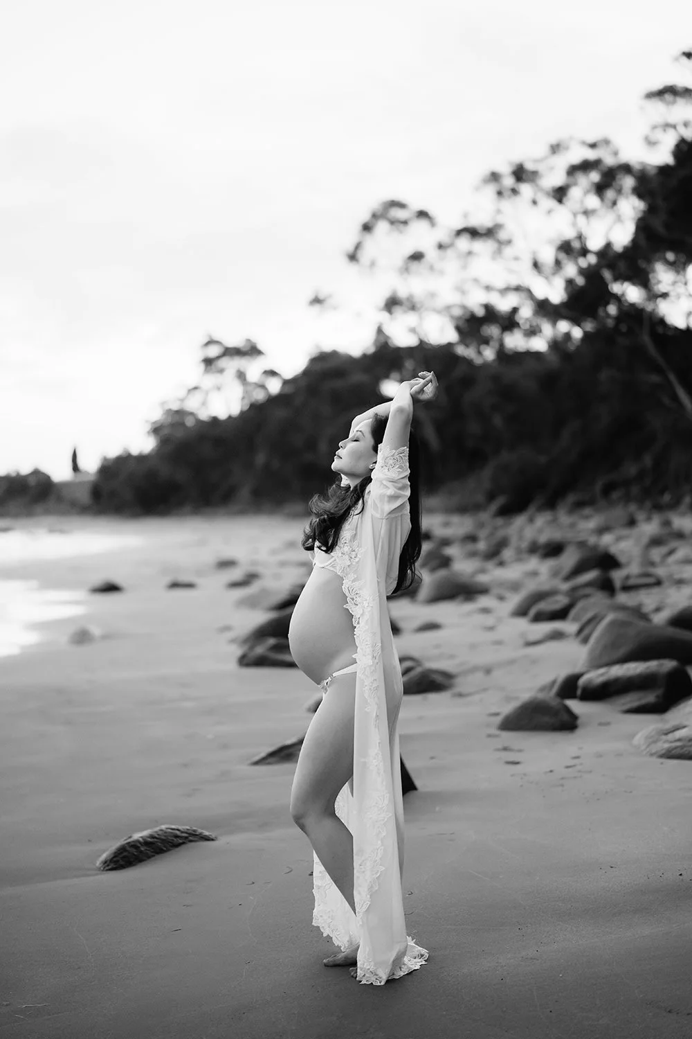 A pregnant woman standing barefoot on a beach with rocks, wearing a sheer lace robe, eyes closed, arms raised, and long hair, with trees and sky in the background.