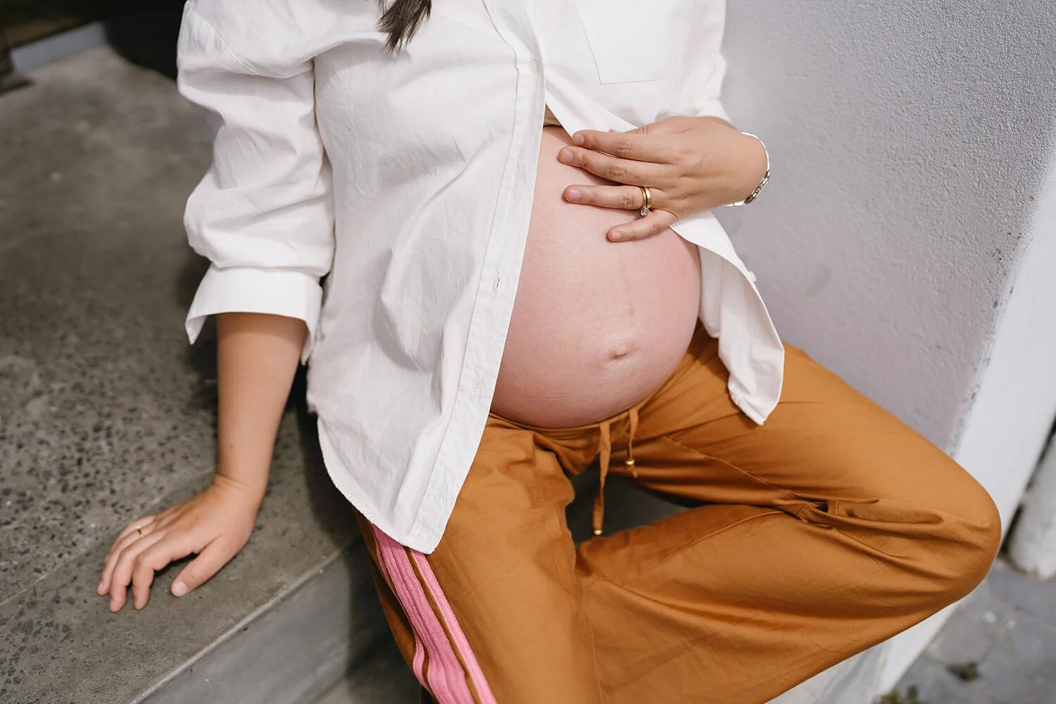Close-up of a pregnant woman sitting on a concrete bench, wearing a white shirt and brown pants with pink stripes, gently holding her belly with one hand, against a white wall.