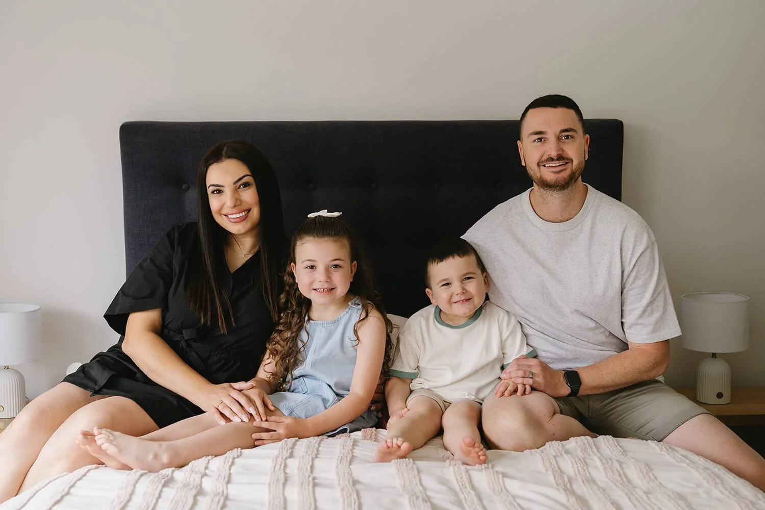 A happy family of four sitting on a bed, smiling at the camera, with a dark headboard and neutral-colored wall behind them. The mother has long dark hair, the father has short dark hair and a beard, and two young children, a girl with curly hair and 