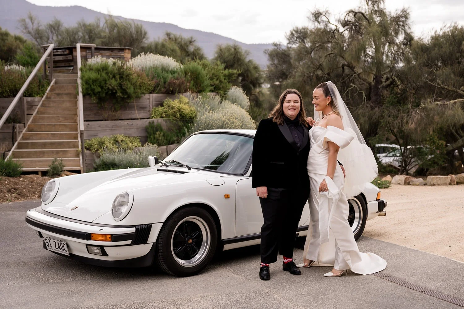 Two women, one in a tuxedo and the other in a wedding dress, standing next to a white classic Porsche 911. The scene is outdoors with a background of trees and a mountain, and they are smiling at each other.