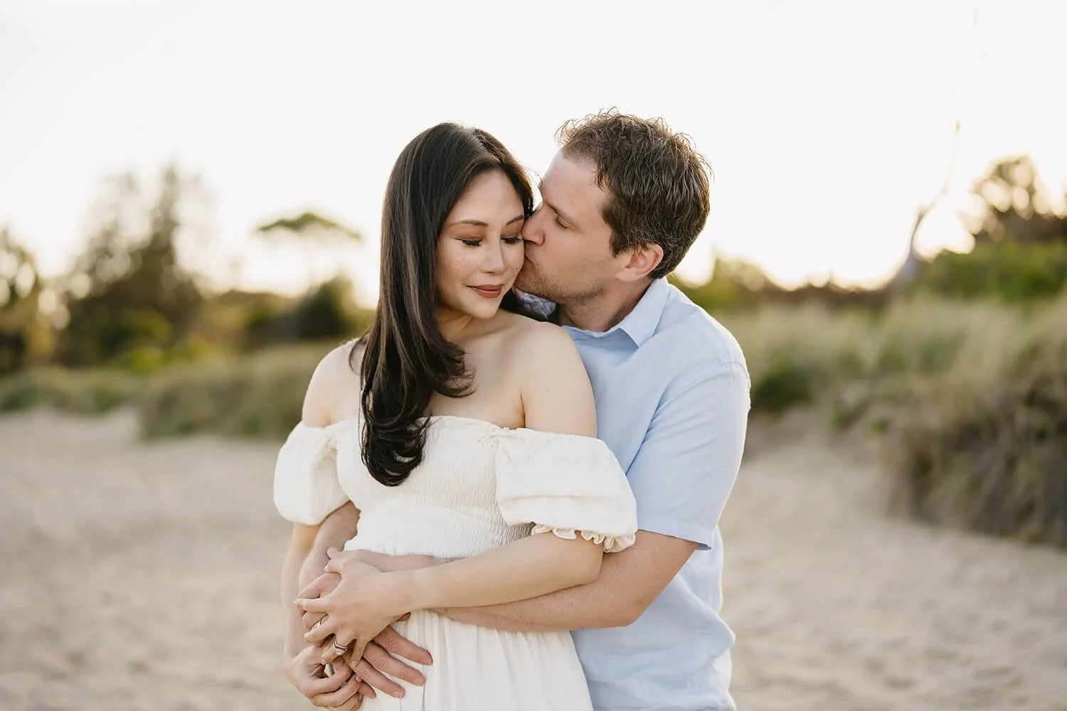 A couple standing on a sandy outdoor path, embracing and sharing a kiss on the cheek at sunset, with trees blurred in the background.