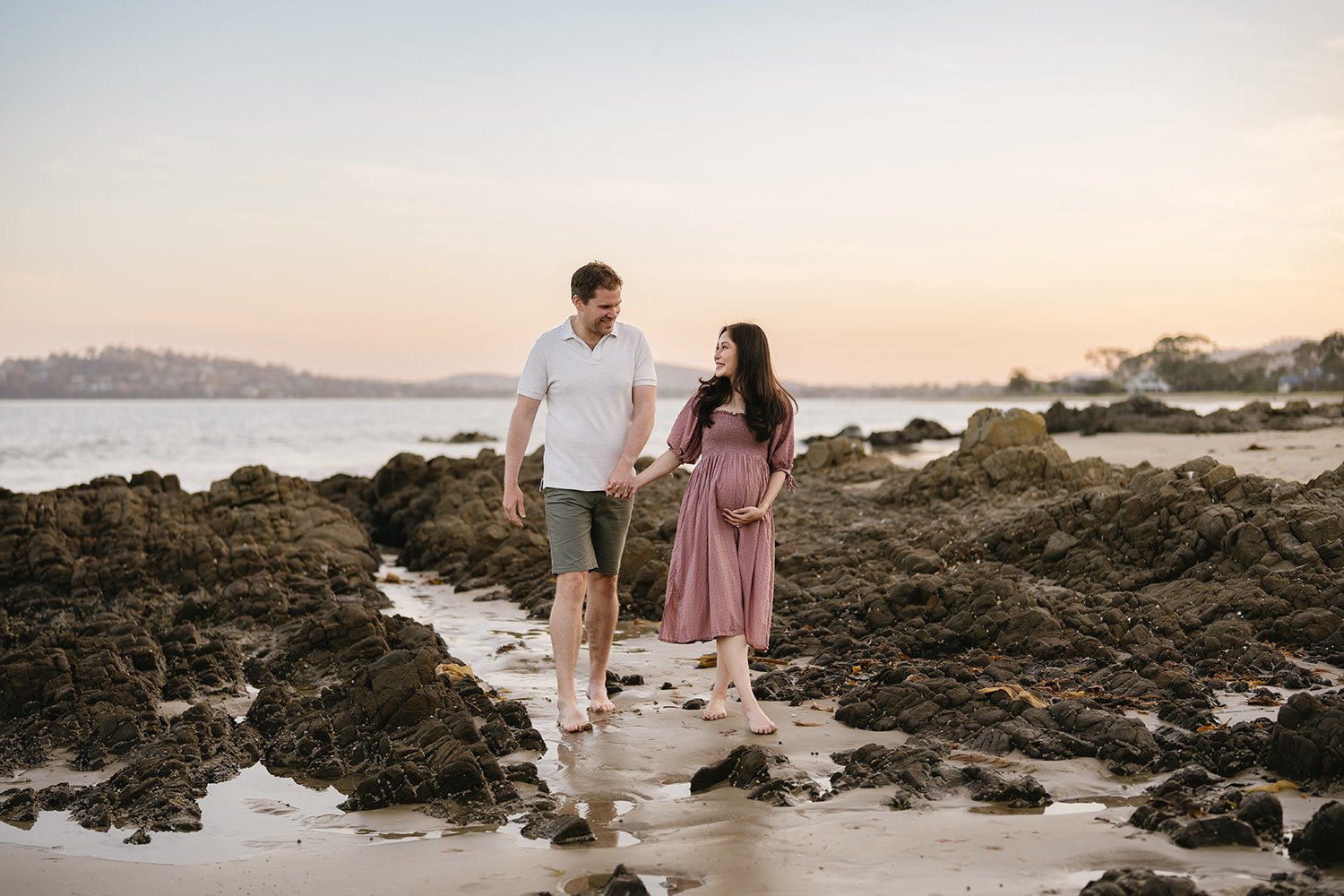 A couple walking hand-in-hand on a rocky beach at sunset. The woman is pregnant and wearing a pink dress, while the man is wearing a white shirt and shorts. They are smiling and looking at each other.
