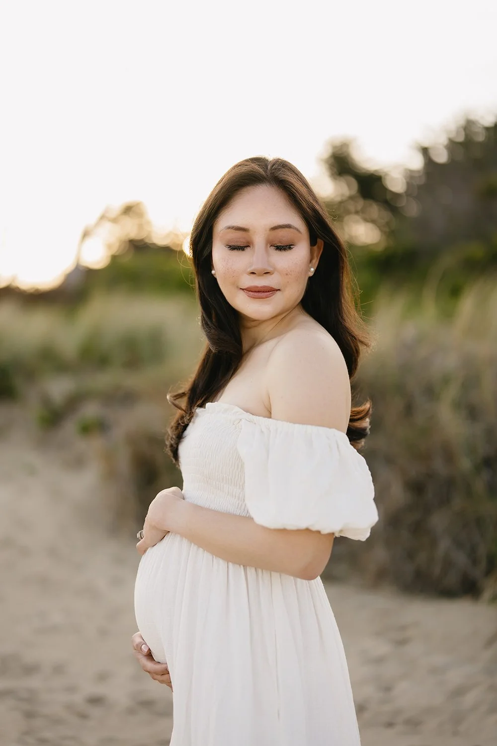 A pregnant woman in a white off-shoulder dress standing outdoors on a beach at sunset, with her eyes closed and a serene expression.