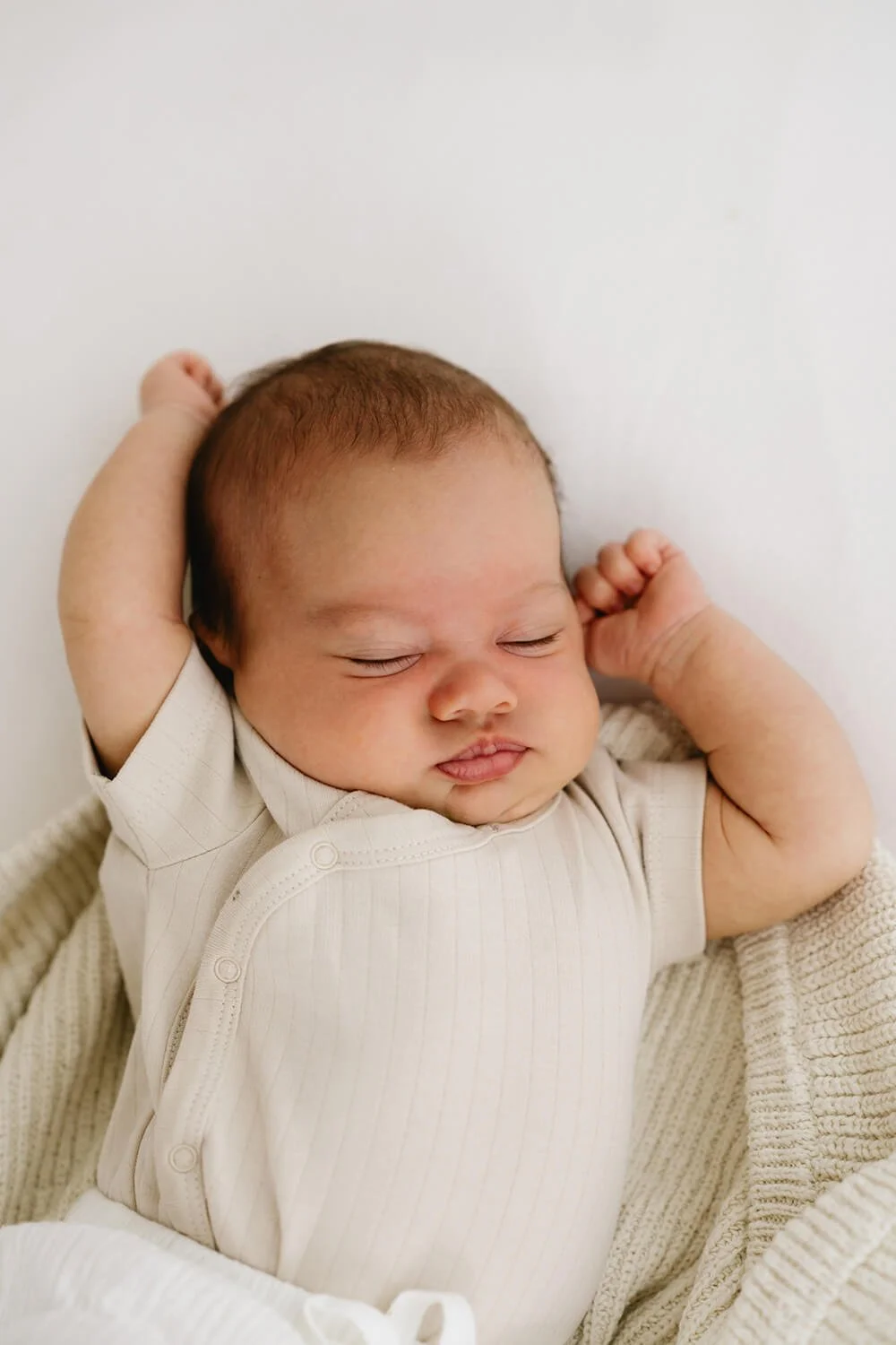 A sleeping baby with arms raised above head, lying on a soft blanket, wearing a light-colored onesie, with a peaceful expression.