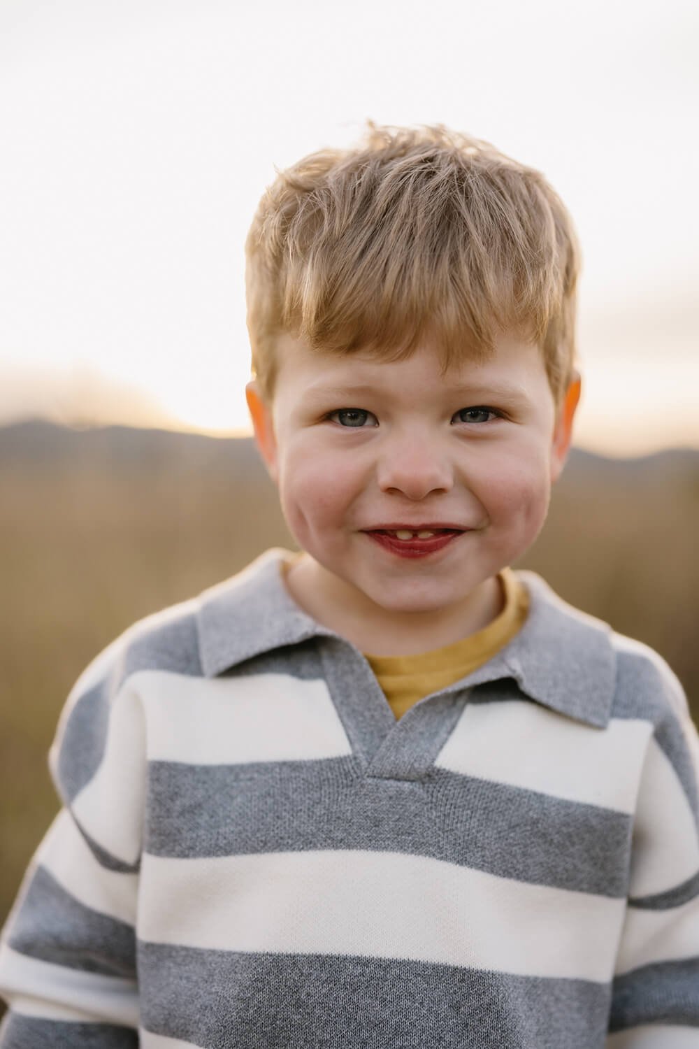 Close-up of a young boy with red hair, blue eyes, and a chipmunk-like face smiling outdoors during sunset.