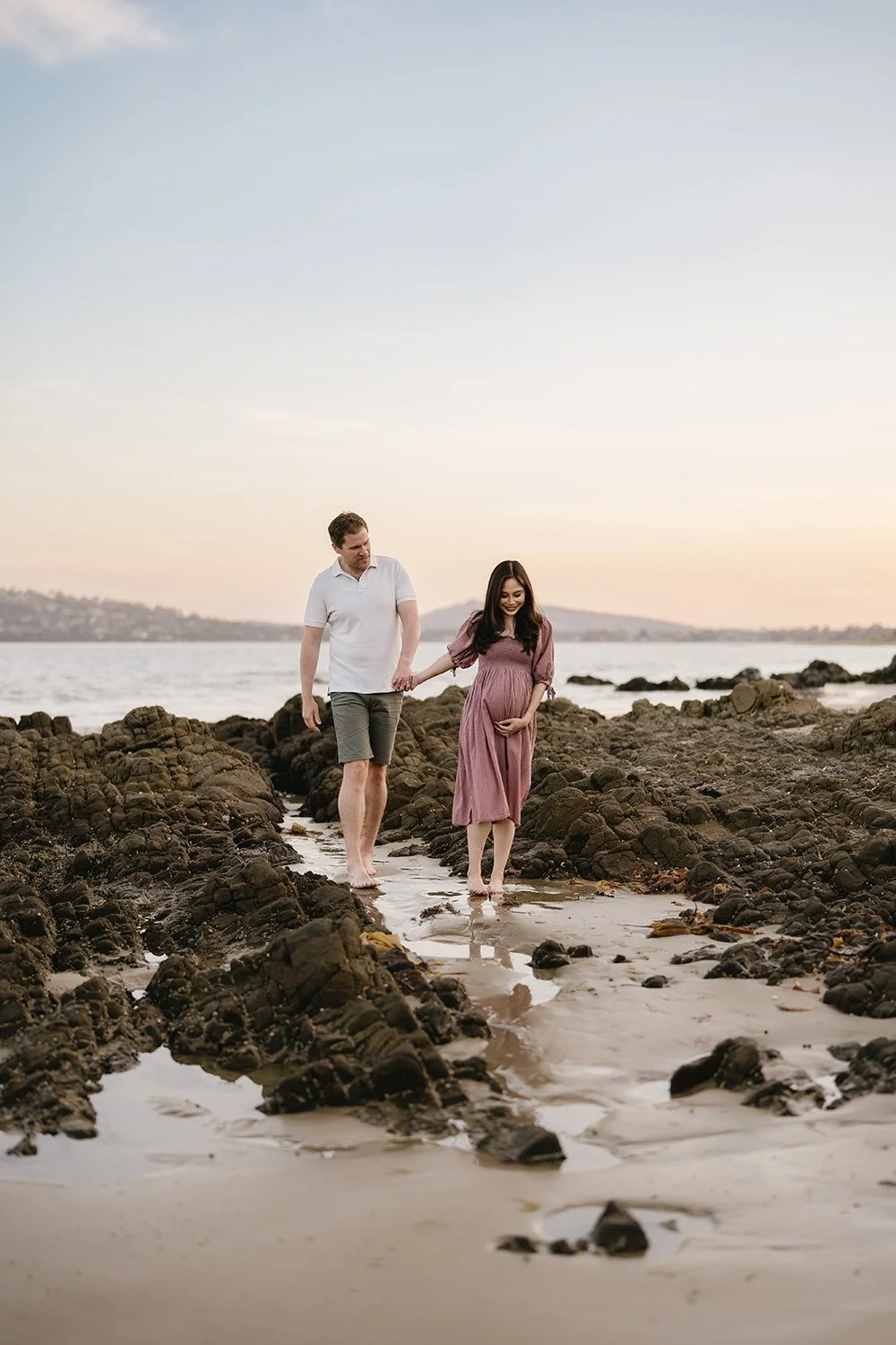 A pregnant woman and her partner walk hand-in-hand along a rocky beach at sunset, with the woman cradling her belly.