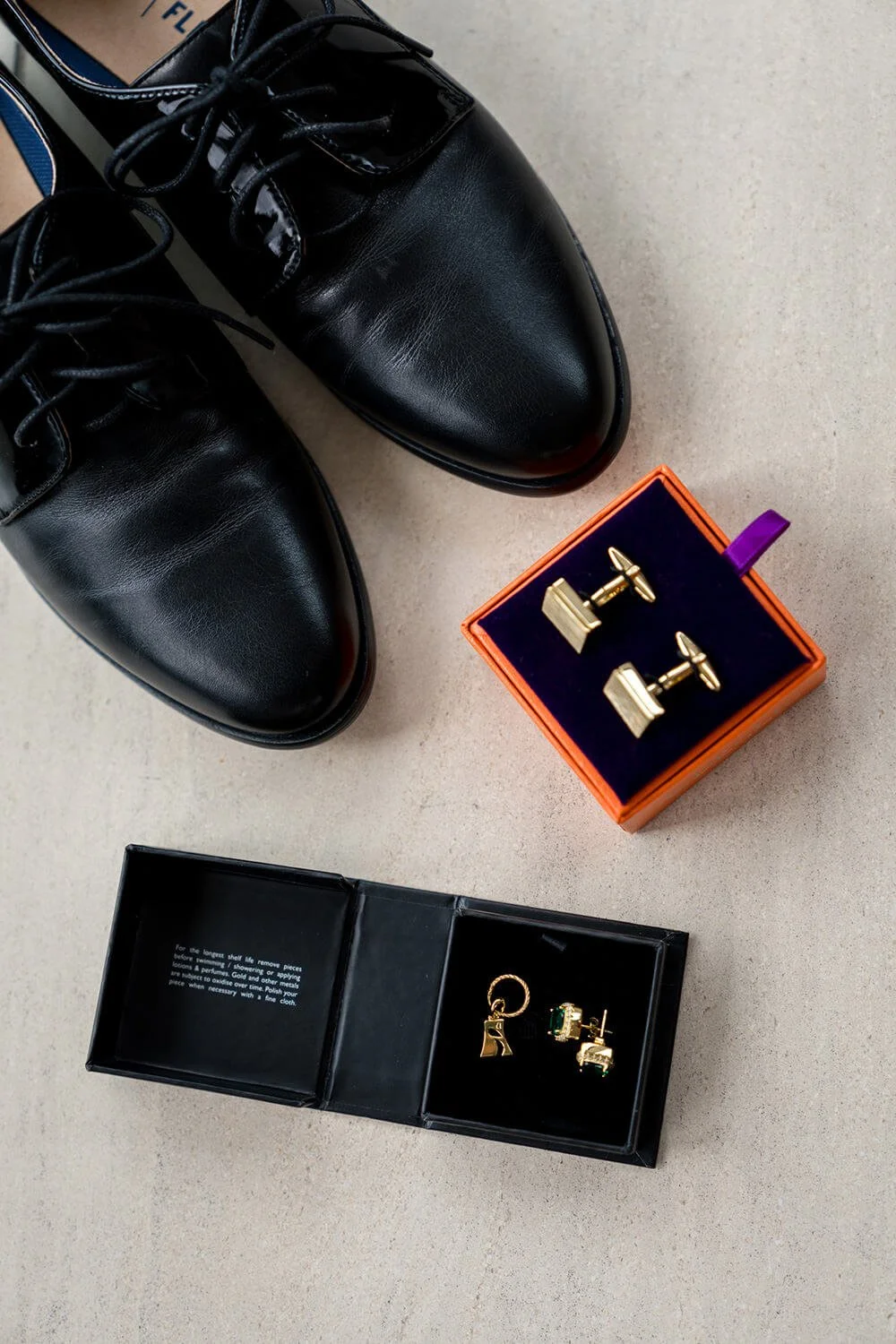 A pair of black leather dress shoes, a box of gold cufflinks with a purple tab, and a jewelry box with gold earrings and a hoop earring inside on a light-colored surface.