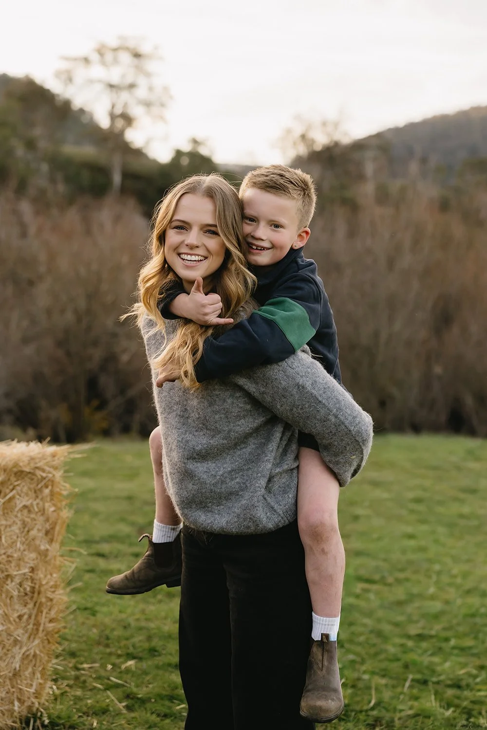 A woman holding a smiling boy on her back outdoors in a grassy area with hay bales and trees in the background.