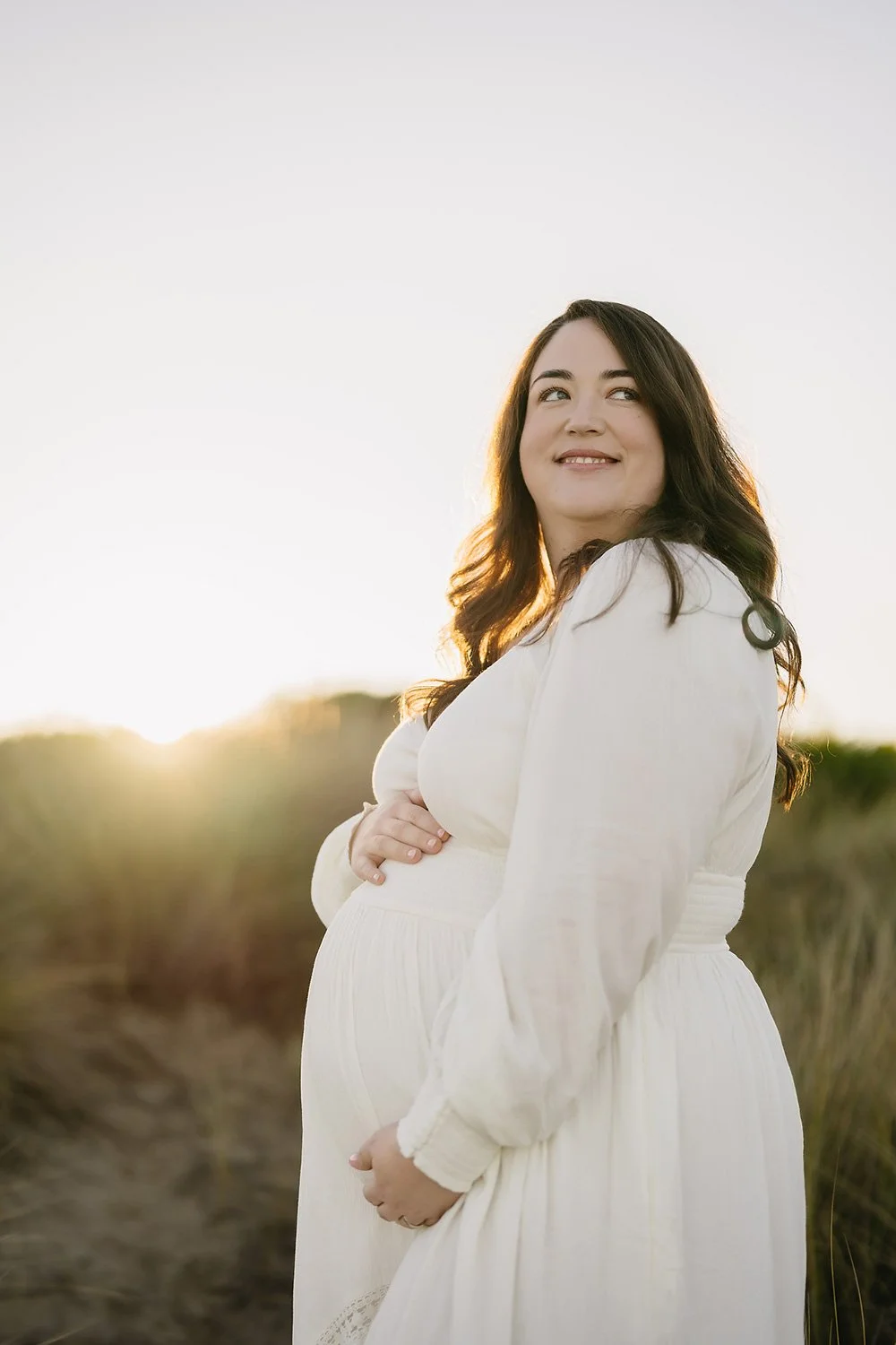 A pregnant woman in a white dress standing outdoors at sunset, smiling and looking up.