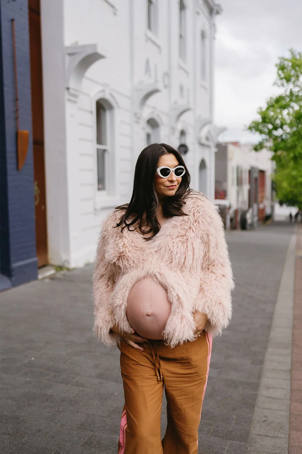 A pregnant woman wearing a fluffy pink jacket, orange pants with pink stripes, and white sunglasses, standing on a city sidewalk with white buildings and green trees in the background.