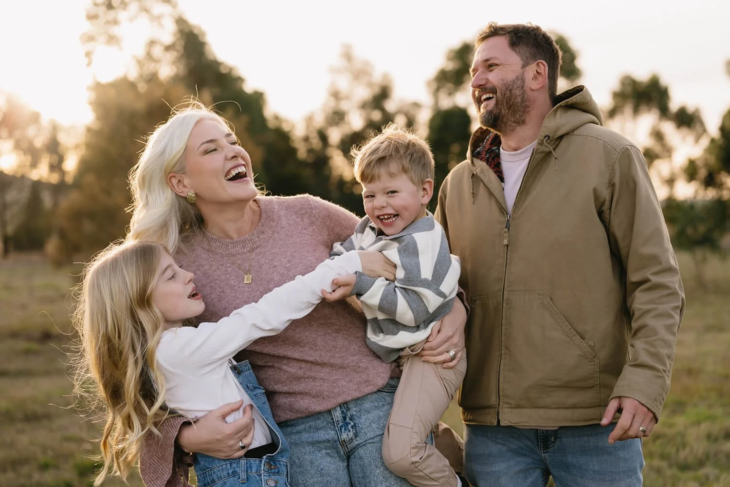 A happy family of four laughing outdoors in a field at sunset. The mother, with blonde hair, is holding her young son, who is grabbing her arm and smiling. The father, with a beard, is standing nearby, smiling as he looks at the family. The daughter 