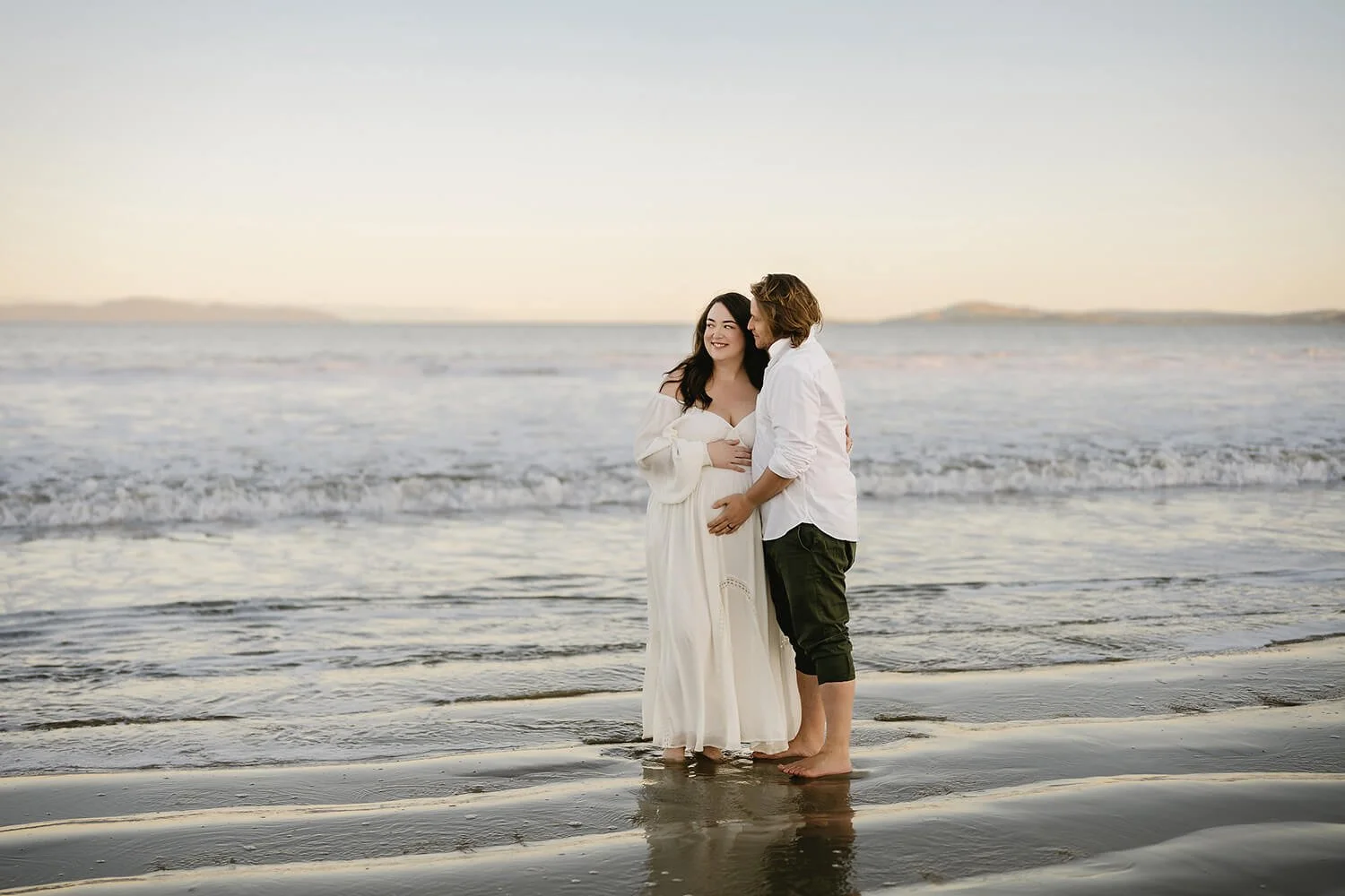 A pregnant woman and a man standing barefoot on the beach, smiling and holding each other during sunset.