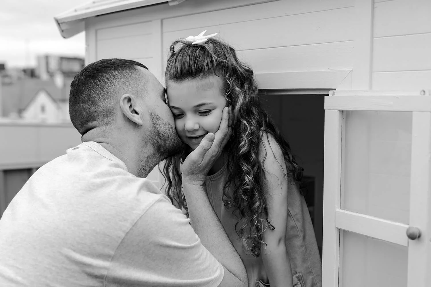 A man is kissing a young girl on the cheek, holding her face tenderly. The girl, with long curly hair and a bow in her hair, is smiling and leaning into the man's touch. They are near a small structure, possibly a playhouse, with a neighborhood in th