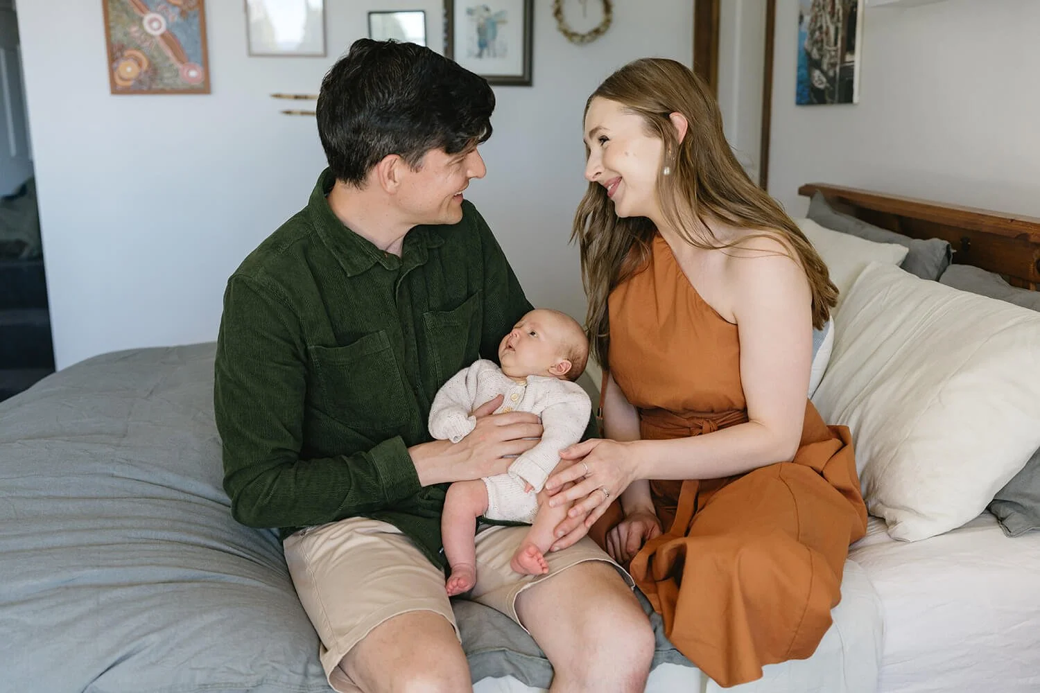 A young family sitting on a bed, with a man holding a baby and a woman reaching out to touch the baby's hand, all smiling and looking at each other.