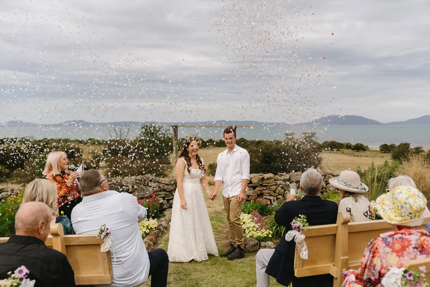 A wedding ceremony outdoors with a couple holding hands and smiling, surrounded by seated guests in a garden with a stone wall, overlooking a body of water and mountains, with confetti in the air.