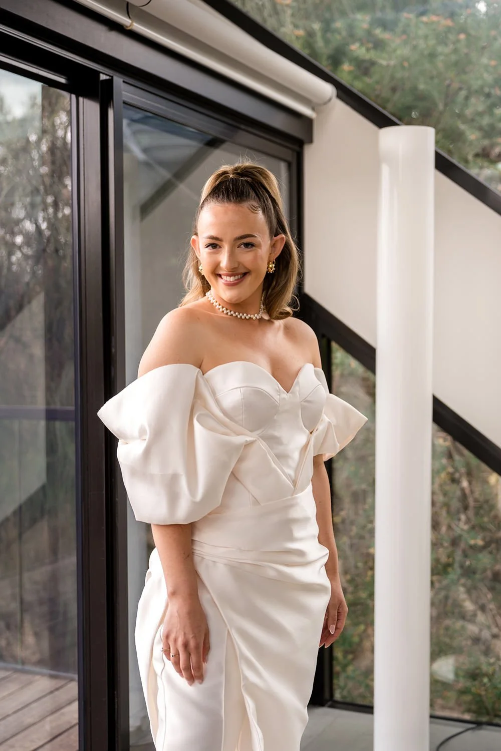 A woman in an elegant white off-shoulder gown with puffy sleeves, standing indoors near a glass sliding door, smiling warmly. She is wearing pearl jewelry and has styled her hair in a high ponytail.