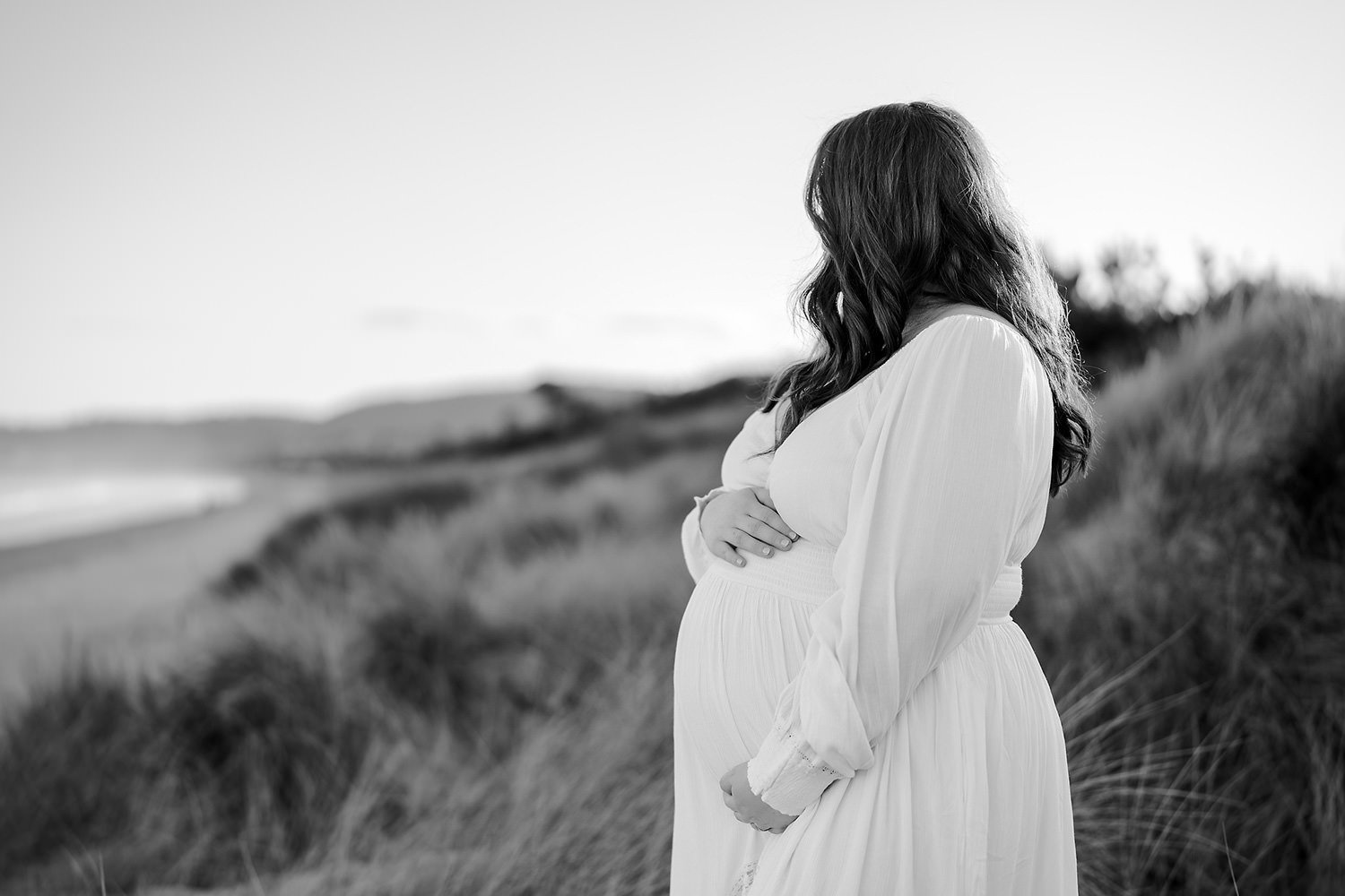 Black and white photo of a pregnant woman in a flowing dress standing outdoors on a grassy hill near the beach, looking away from the camera.