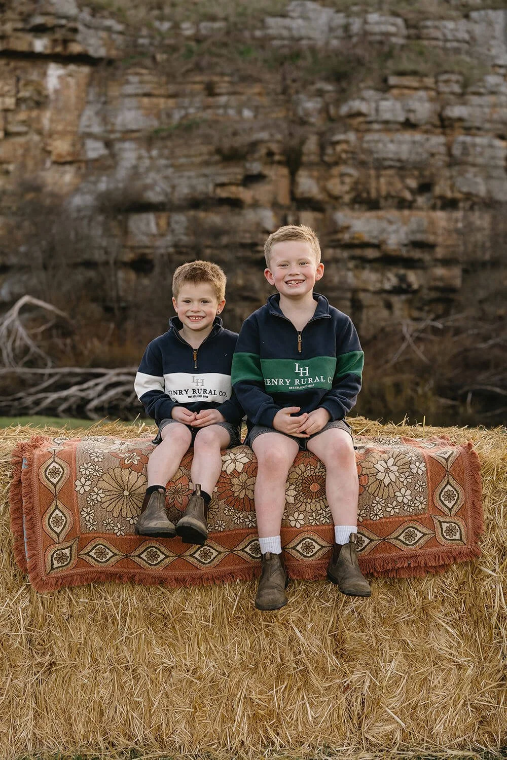 Two boys sitting on a blanket on top of a hay bale outdoors, with a rocky background. Both are smiling and wearing matching jackets with the logo 'Henry Rural Co.'.