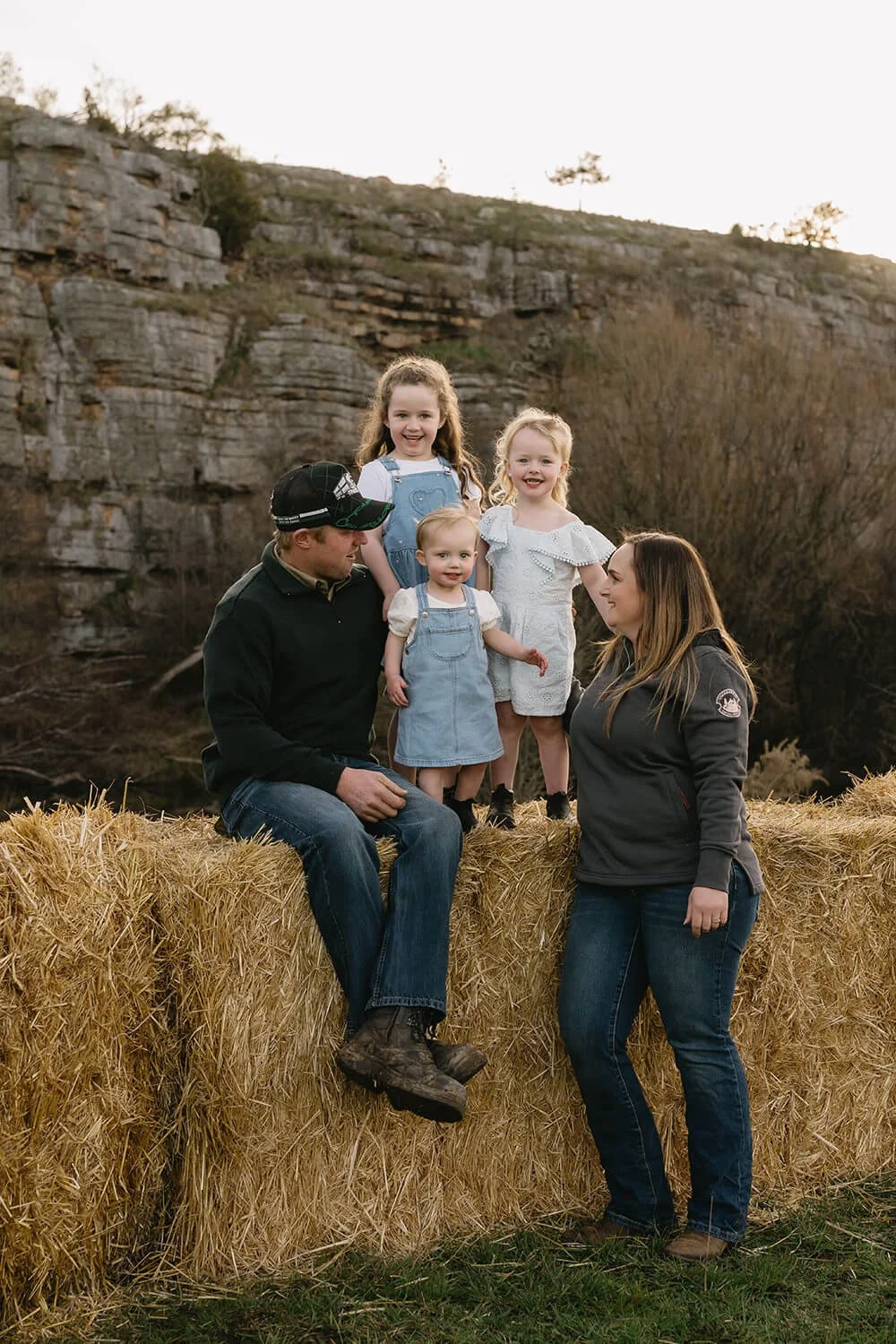 A family of five with three young girls sitting and standing on a hay bale outdoors with rocky cliffs and trees in the background during sunset.