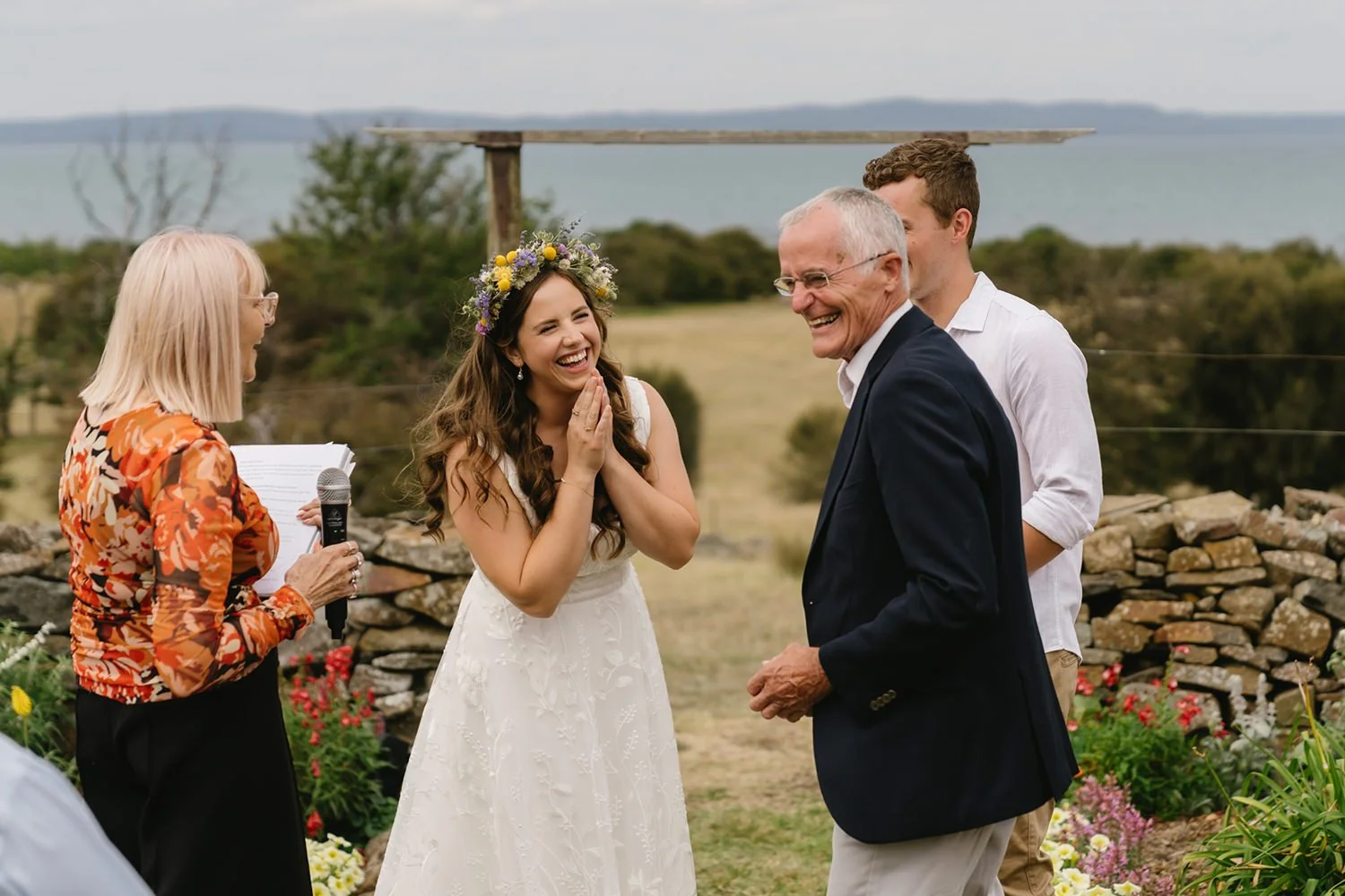 A wedding ceremony outdoors with a bride in a white dress and floral crown, a groom in a dark blazer, and a woman officiant holding a microphone, all smiling and laughing.