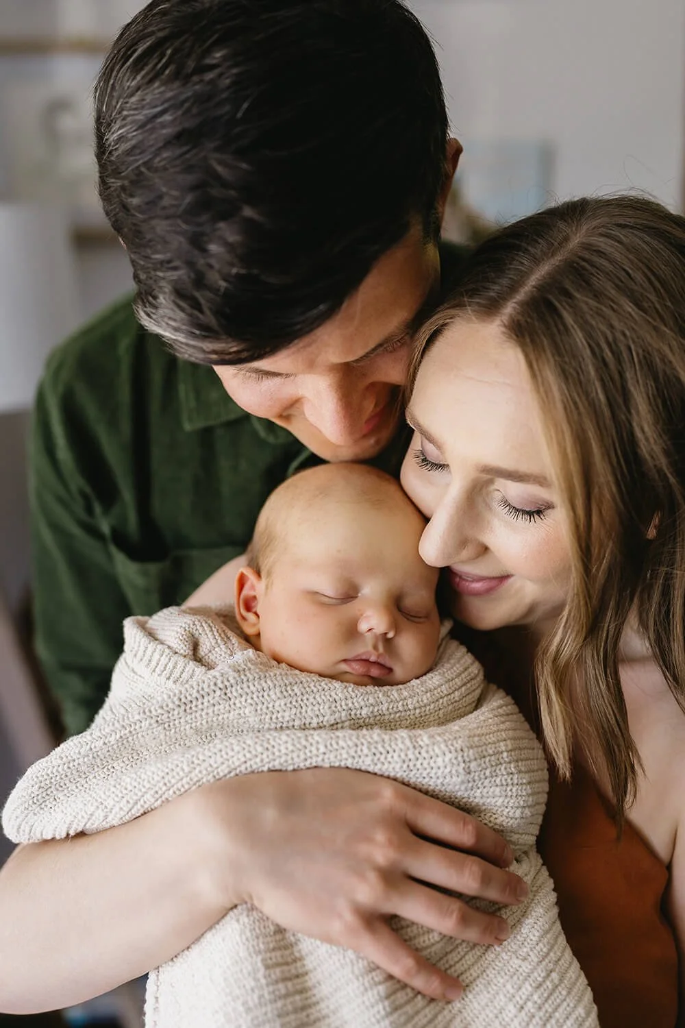 A young mother and father hold their sleeping newborn wrapped in a beige blanket, sharing a tender moment in a cozy indoor setting.