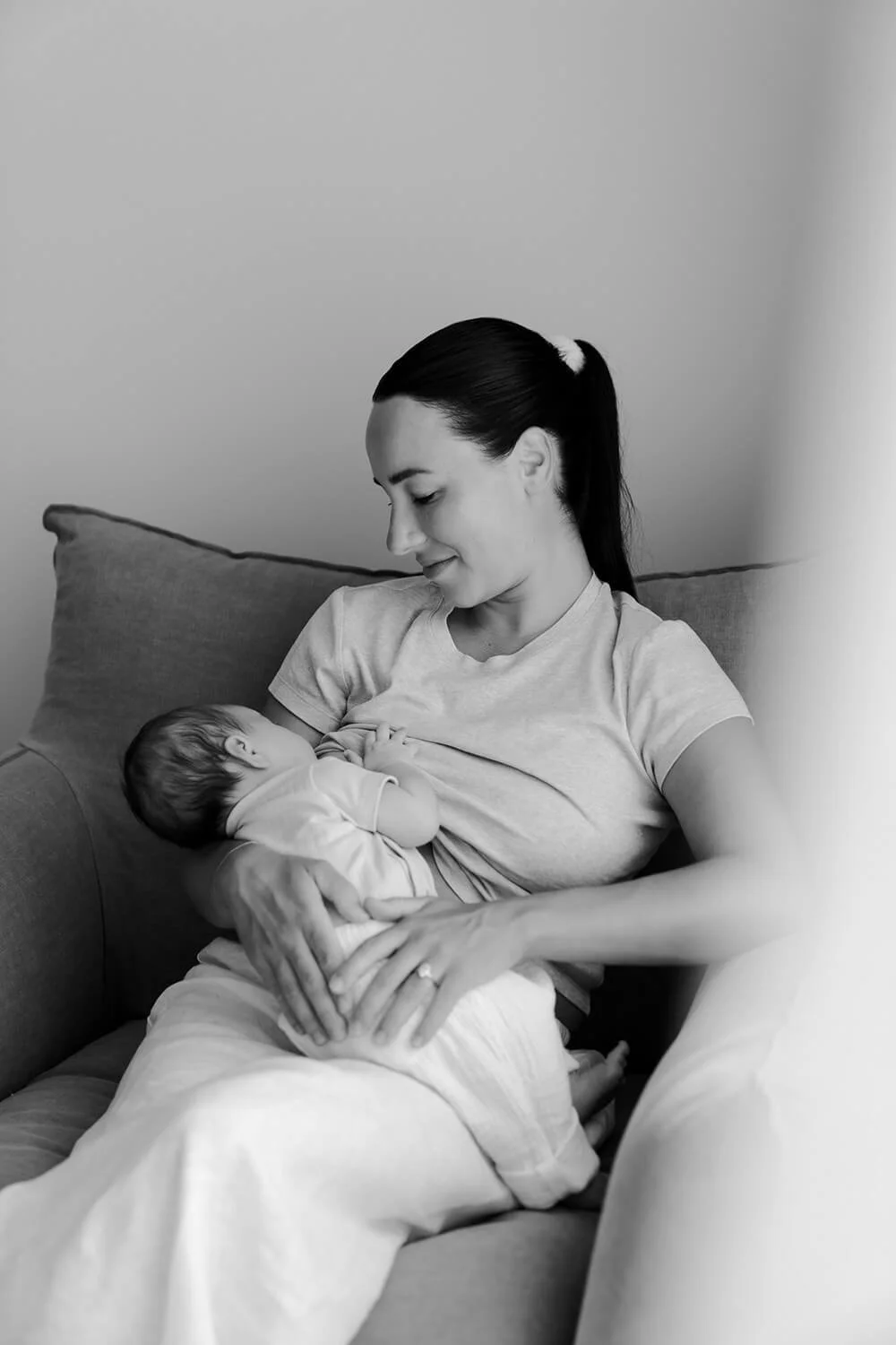 A woman breastfeeds her newborn baby while sitting on a couch in a black and white photo.