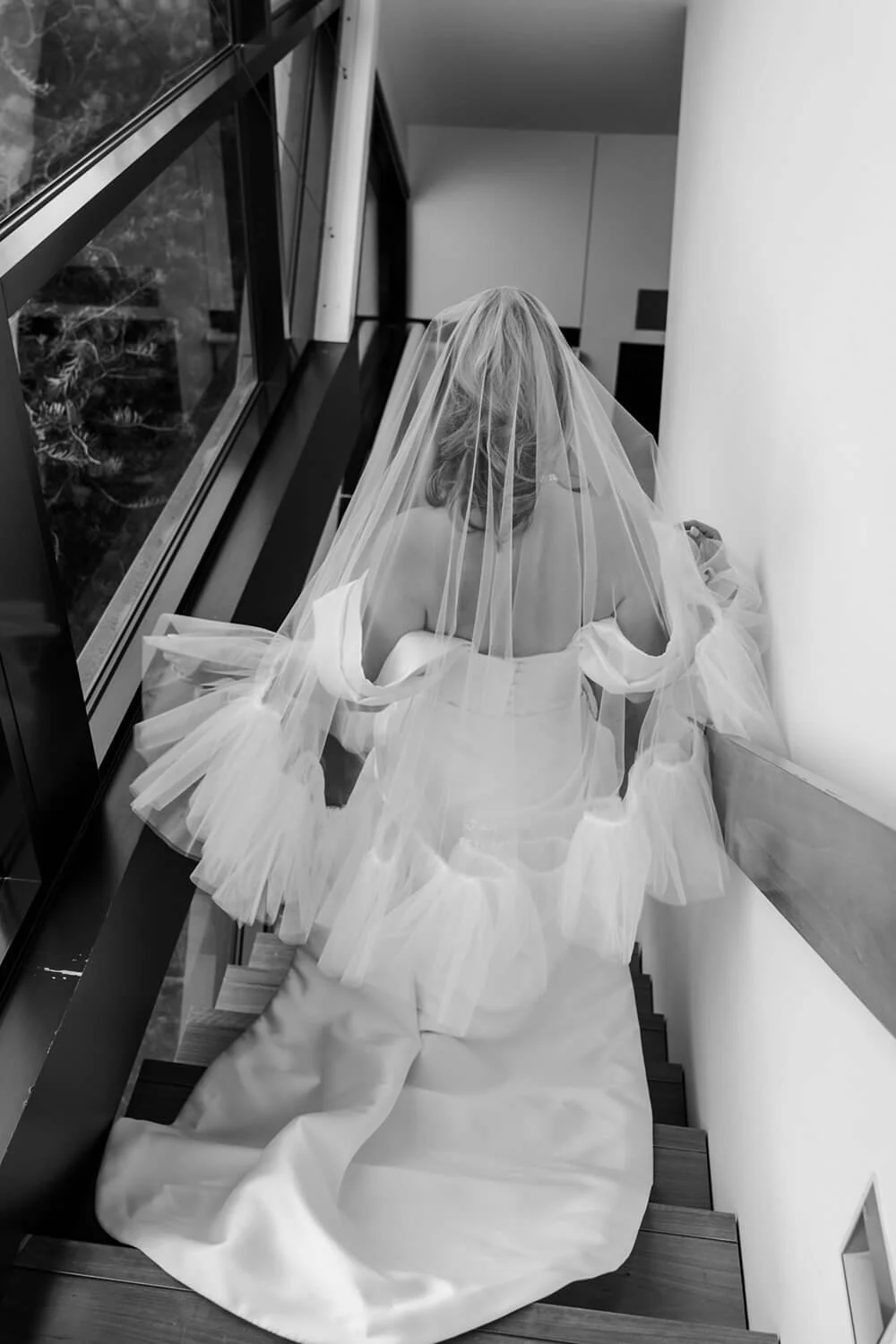 Bride with veil descending wedding dress on staircase near a large window.