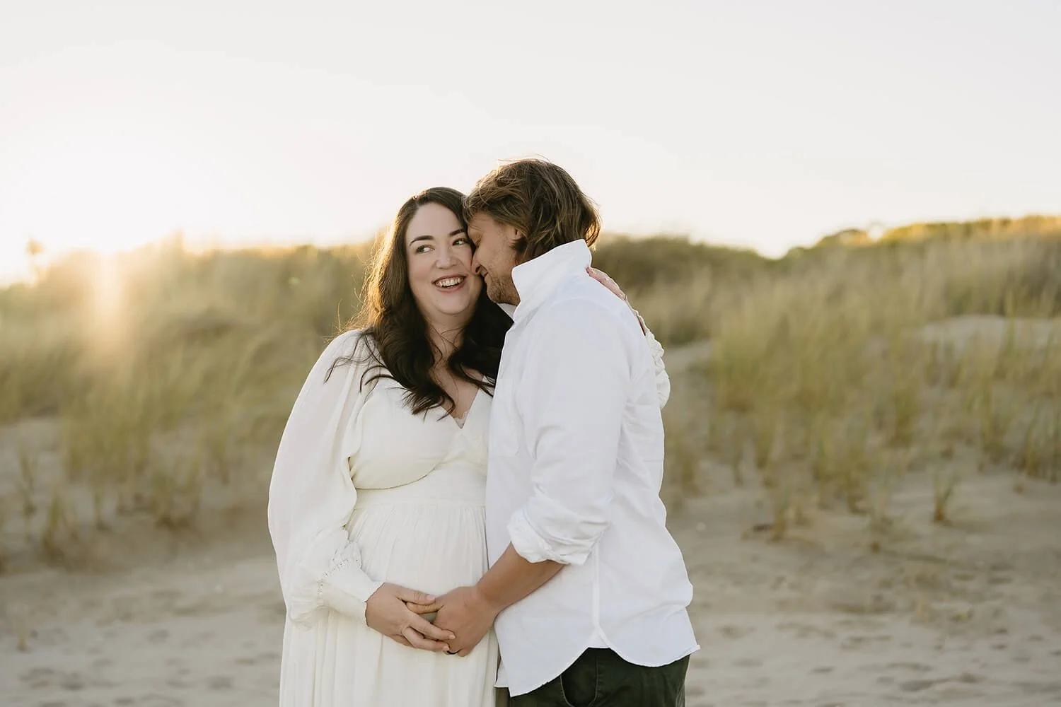 Couple standing on the beach during sunset, with the woman visibly pregnant, sharing an intimate moment.