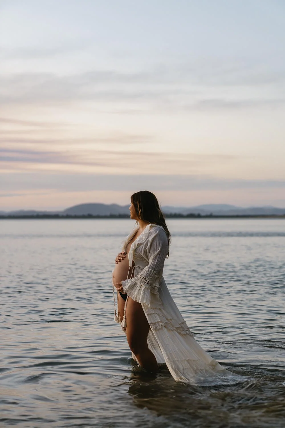 Pregnant women in robe standing in the water showing her pregnant belly being photographed by maternity photographer