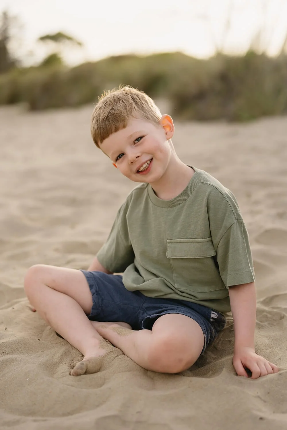 A young boy with short light brown hair smiling while sitting on the sandy beach at Roches Beach Hobart.