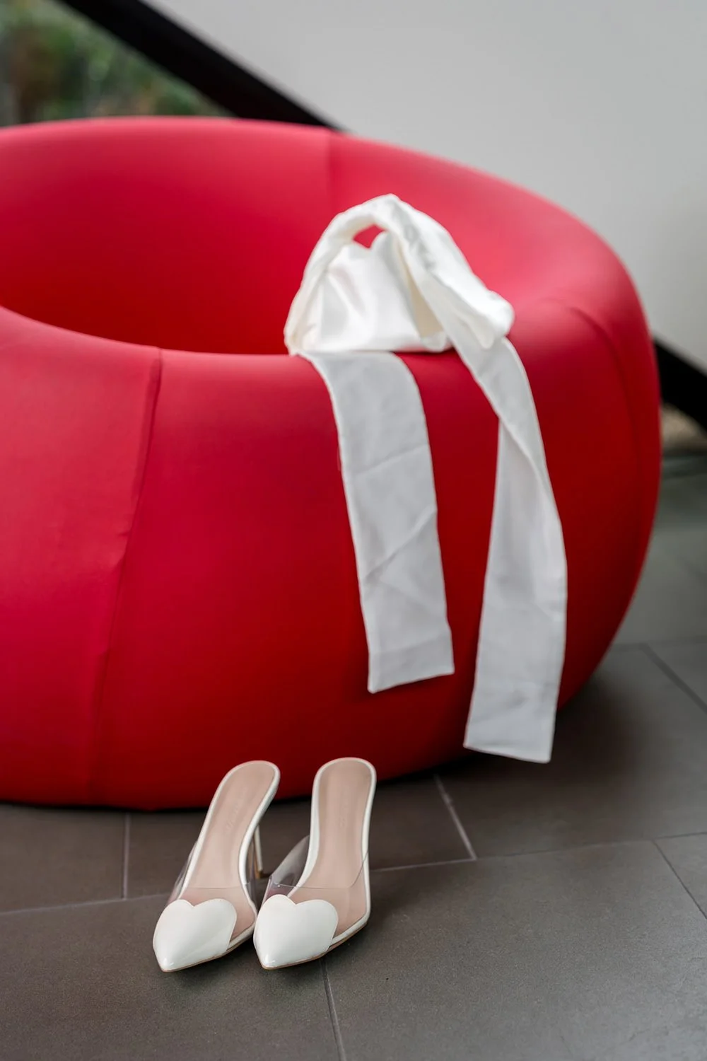 Pair of white high-heeled shoes with heart-shaped toes placed on the floor next to a large red round chair or sofa, with a white cloth draped over its edge.
